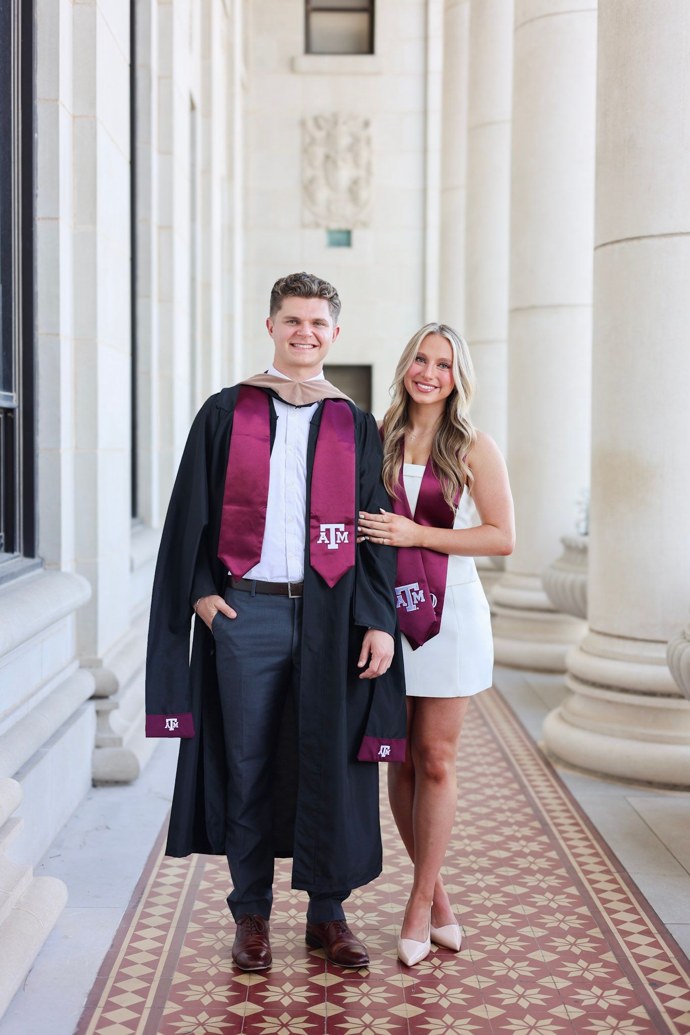 Two Texas A&M graduates in black gowns and maroon stoles posing together in elegant columned corridor.
