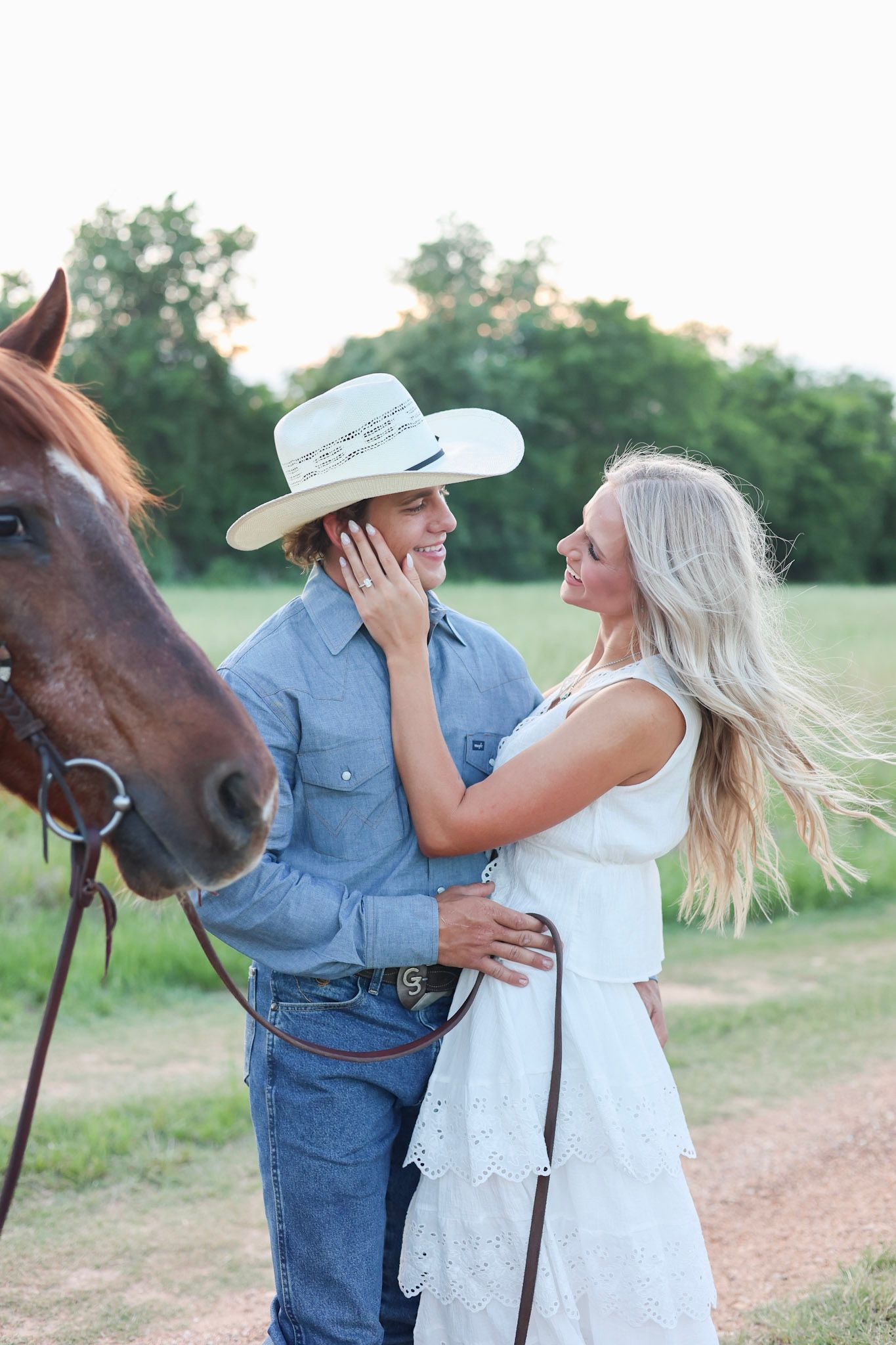 A couple shares a romantic moment near a brown horse, with the man in a cowboy hat and woman in a white dress outdoors.