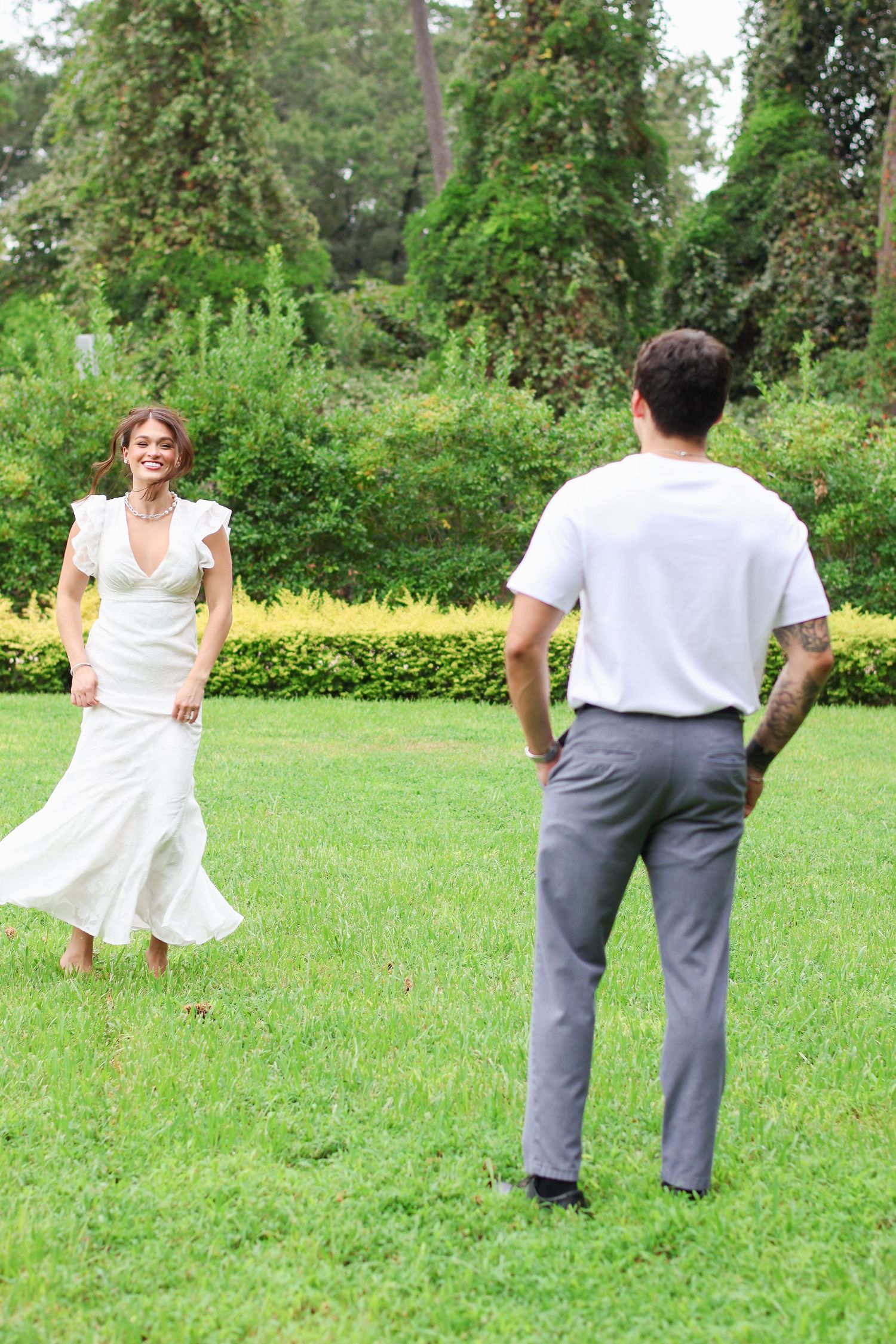 A couple in white and grey clothing dances playfully together on a grassy lawn surrounded by trees.