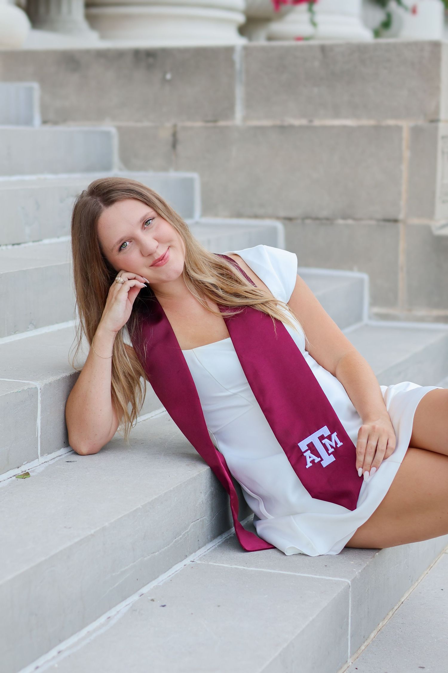 A student in a white dress and maroon Texas A&M stole reclines on concrete steps during a graduation photoshoot.