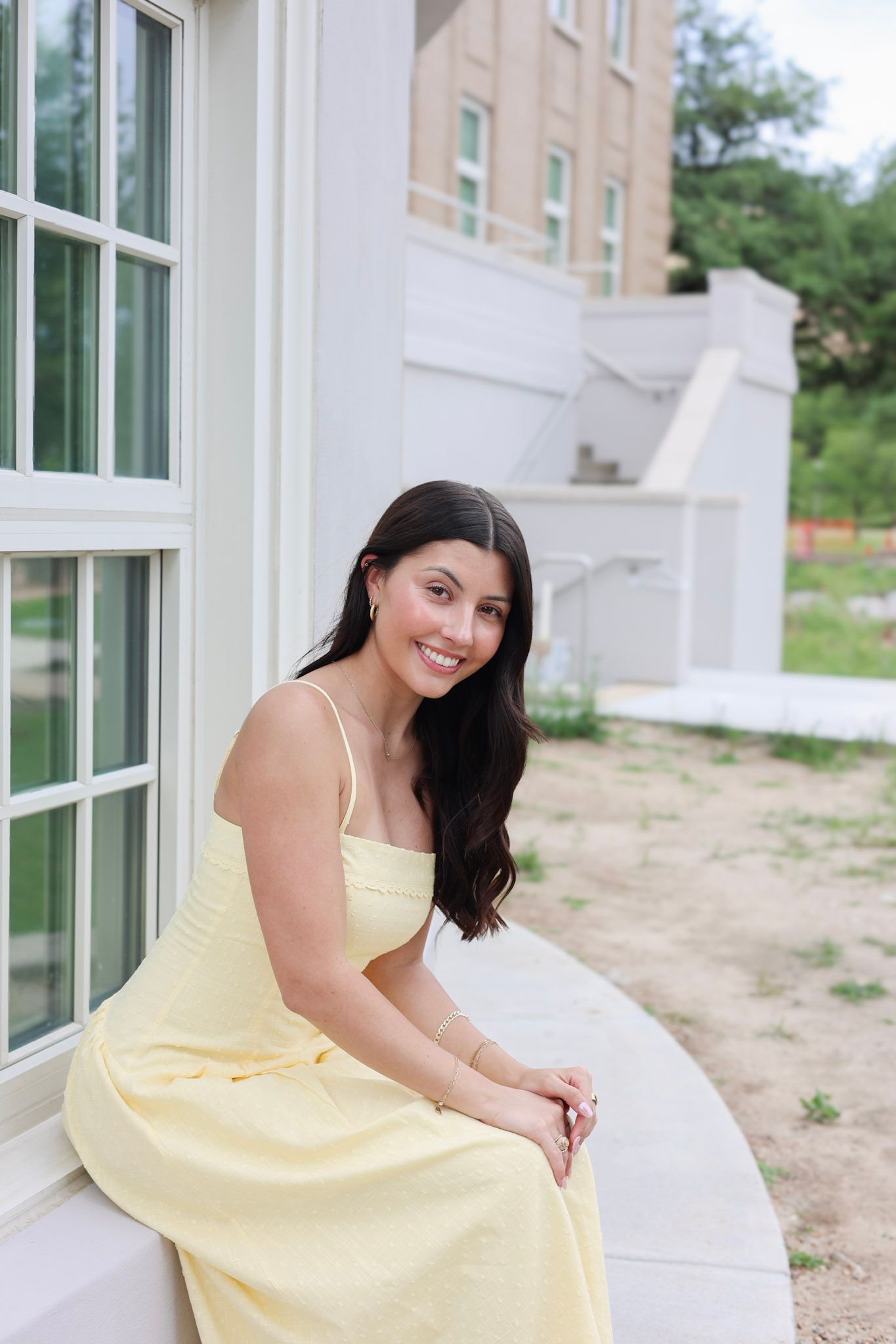 Smiling young woman in yellow sundress sitting on white window ledge outside a building.