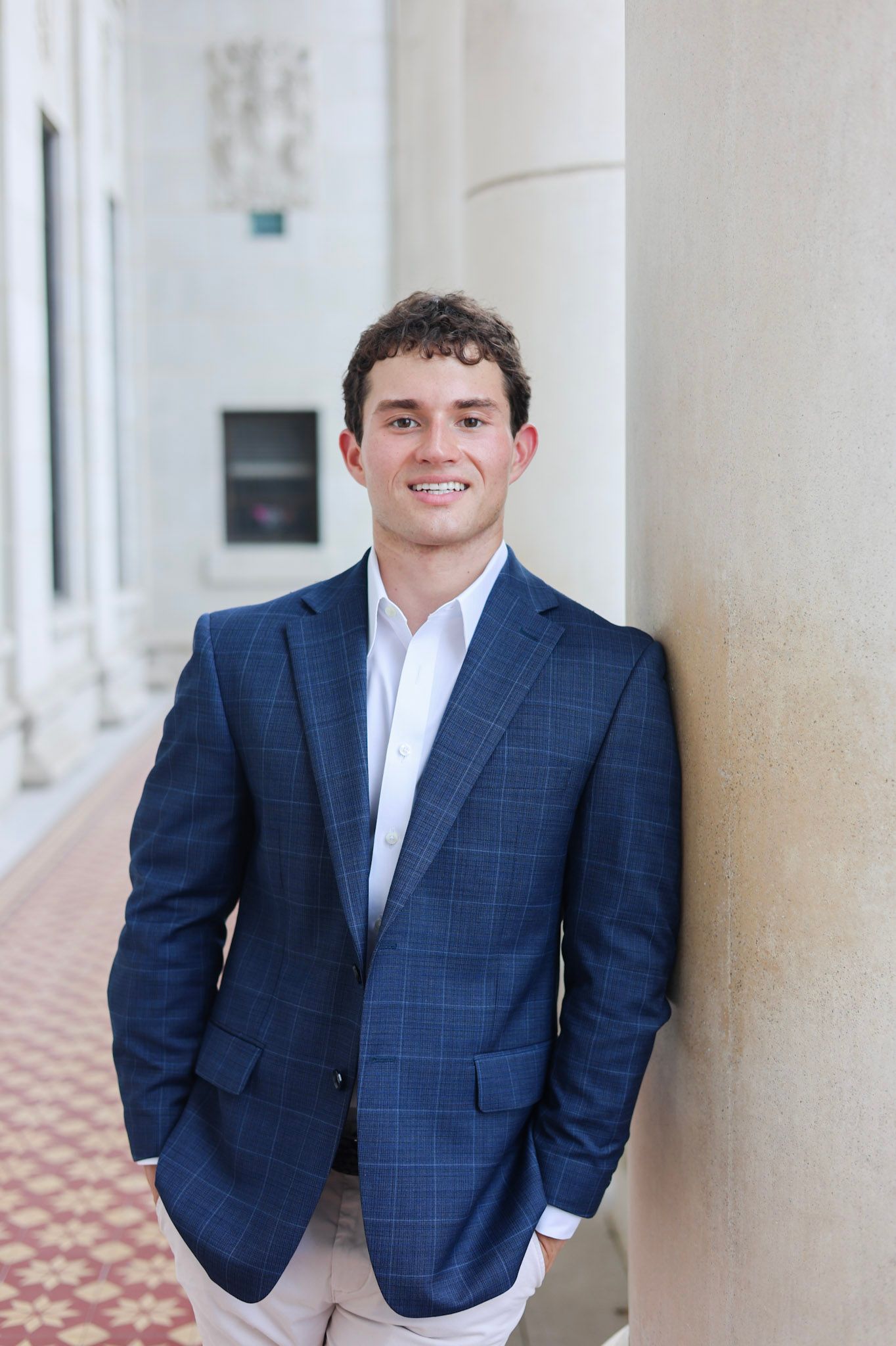 Young man in navy plaid blazer smiling confidently while leaning against a column in an elegant corridor.