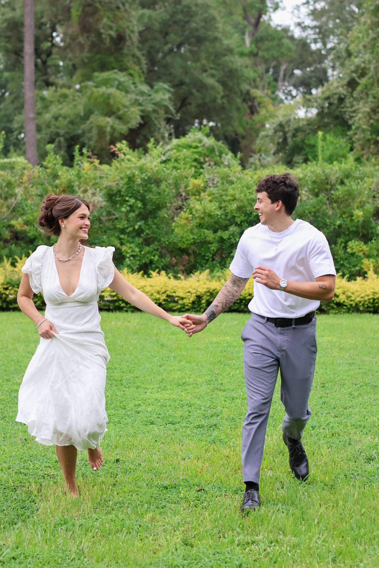 A couple in wedding attire playfully runs and spins across a lush green lawn surrounded by trees and bushes.