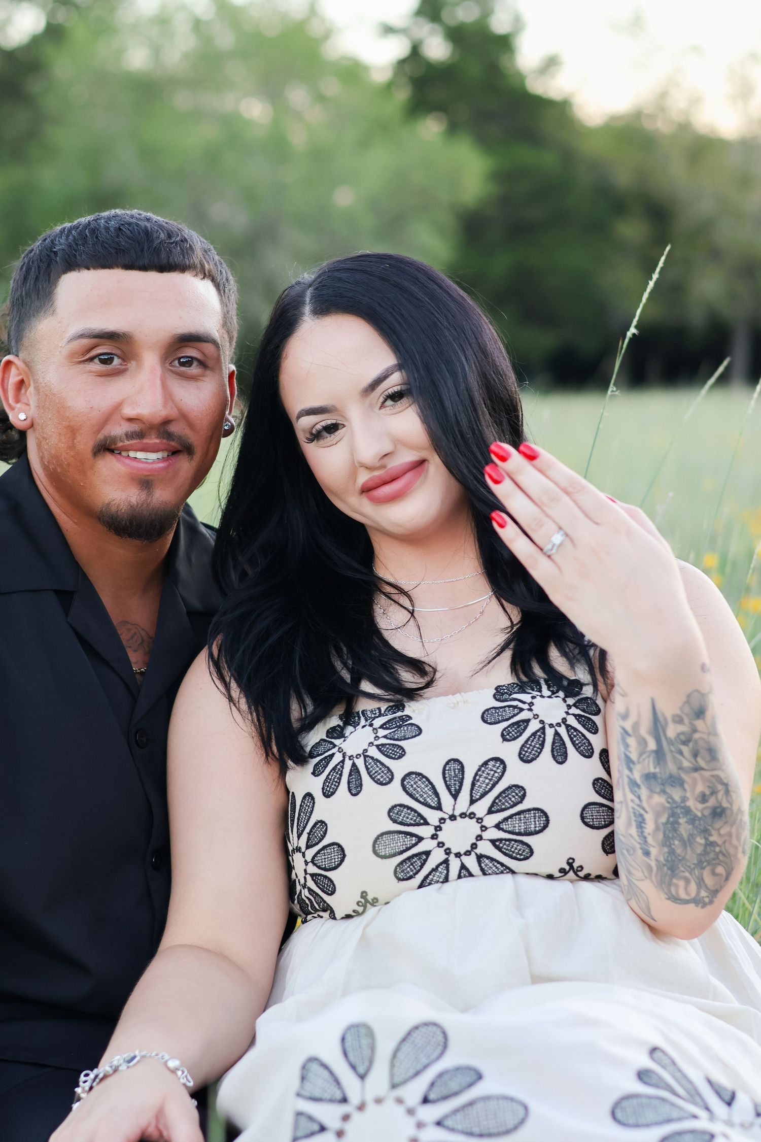 A couple poses together outdoors showing off an engagement ring while wearing coordinating black and floral outfits.