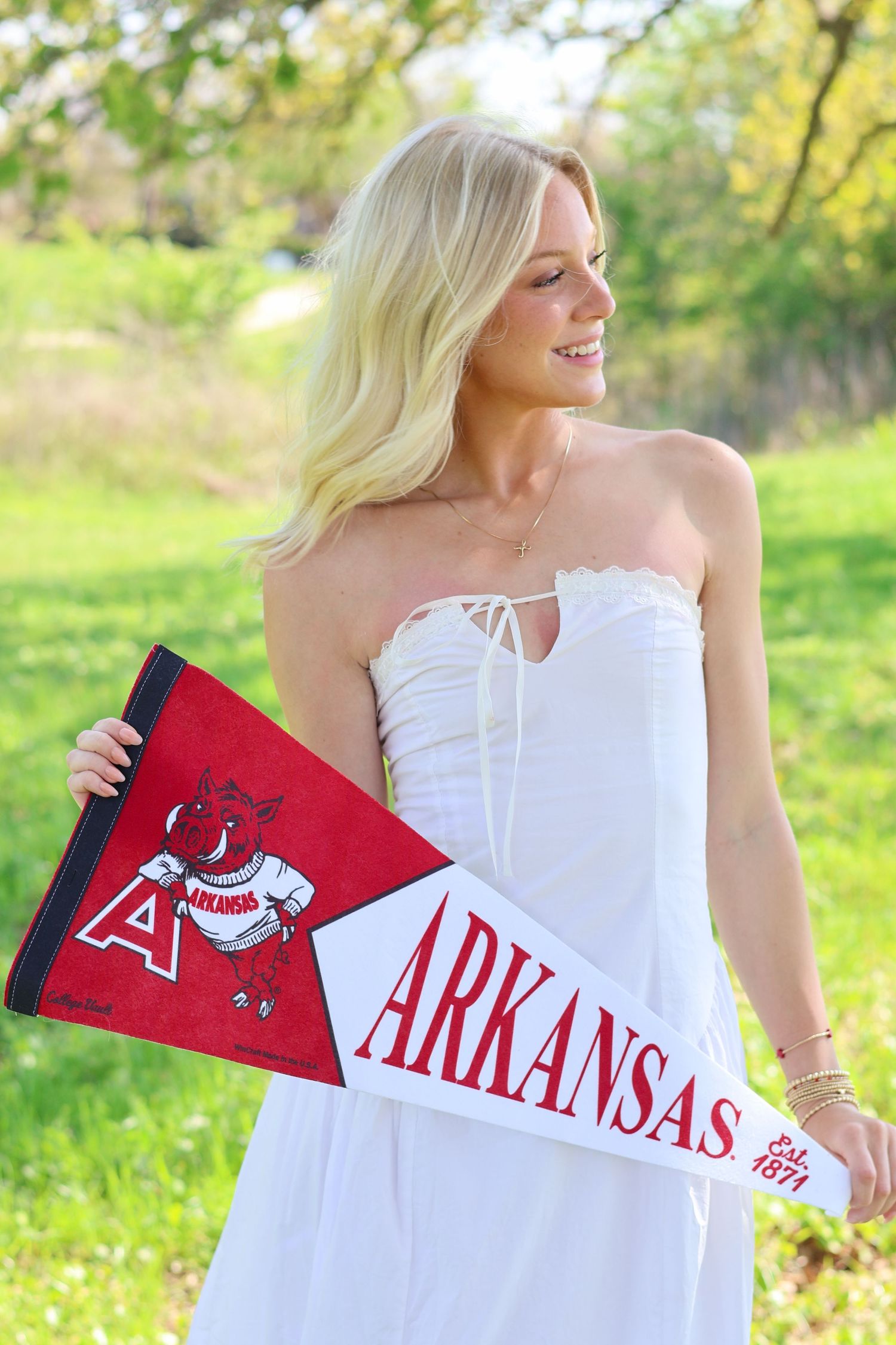Blonde woman in white dress holding Arkansas Razorbacks pennants outdoors.