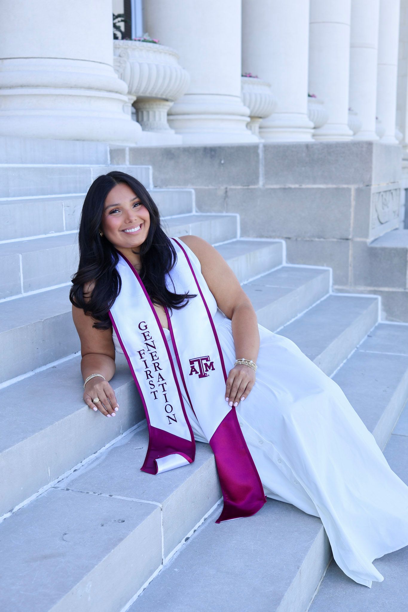 Texas A&M University graduate smiles in white gown and maroon First Generation sash on marble steps.