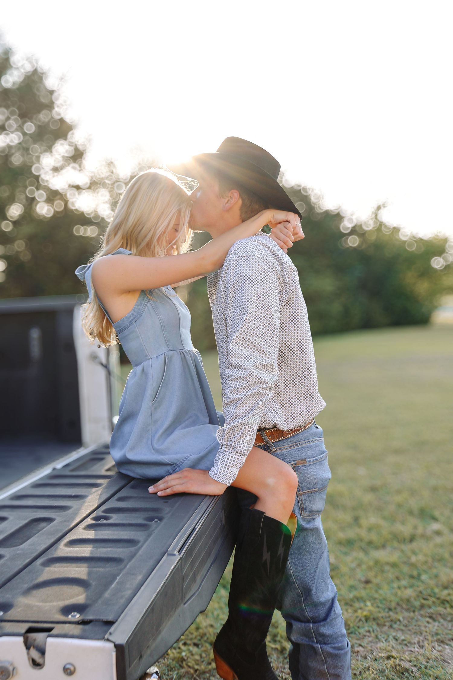 Romantic couple in western wear shares intimate moment on pickup truck tailgate at sunset.