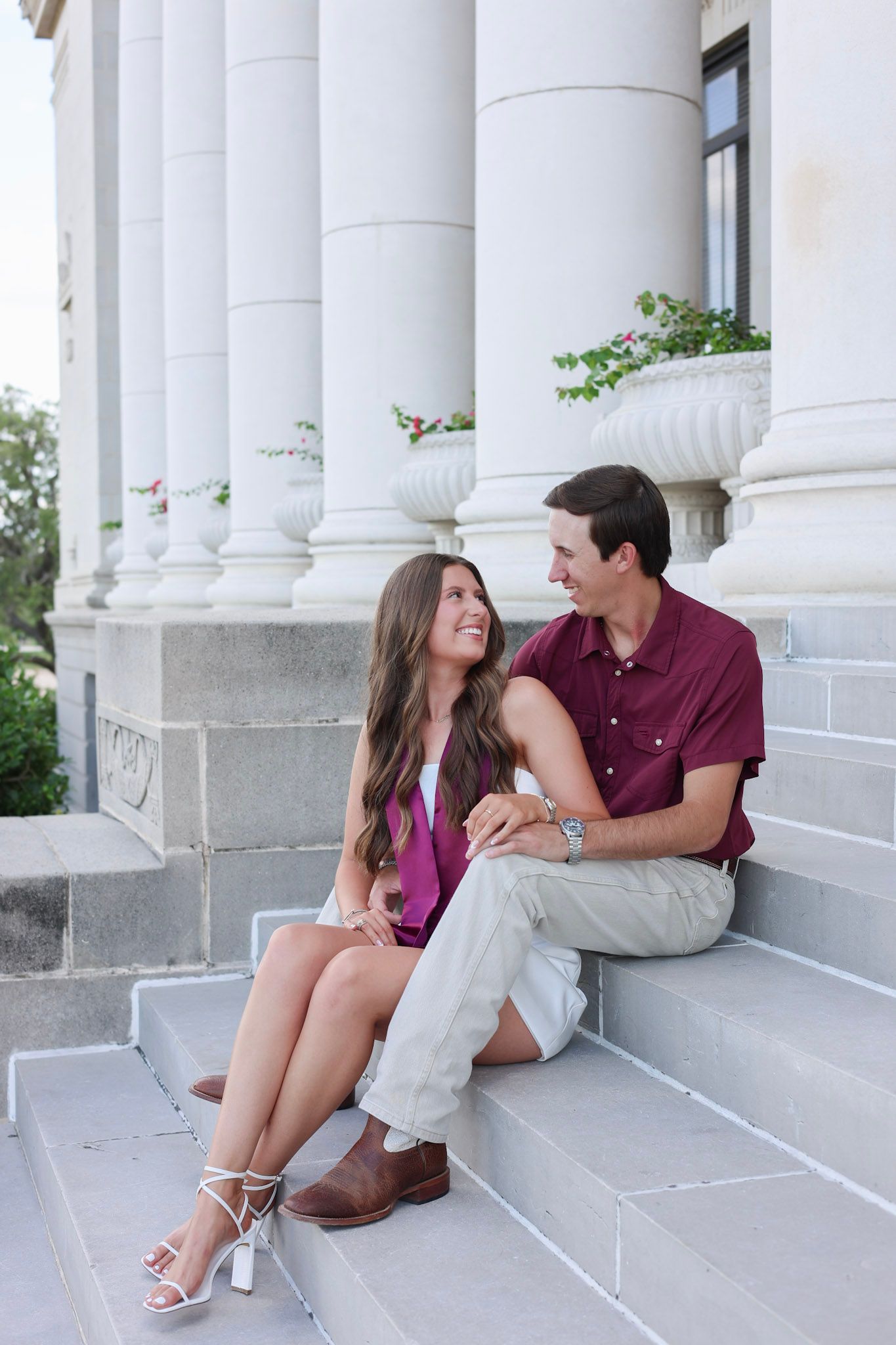 Couple in coordinating maroon and white outfits sit together smiling on steps of columned building.