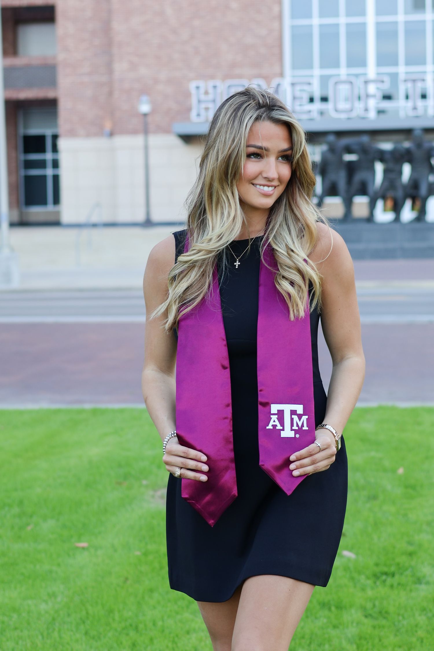 Smiling graduate wearing black dress and maroon Texas A&M stole poses outdoors on campus green.