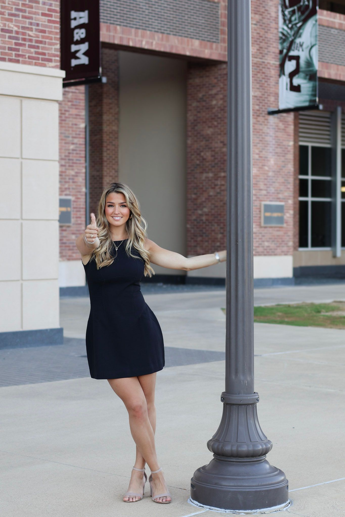 Young woman in black dress giving thumbs up near a lamp post outside Texas A&M University stadium.