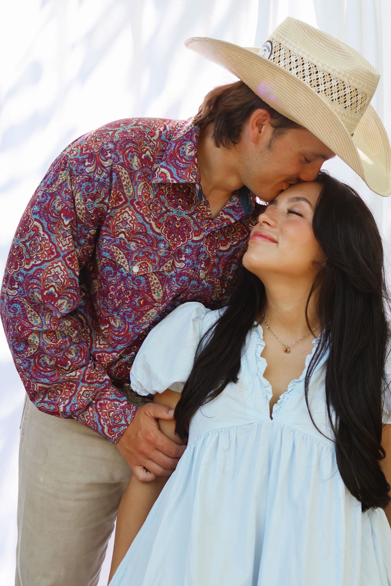Romantic couple portrait with cowboy hat and paisley shirt against white background.