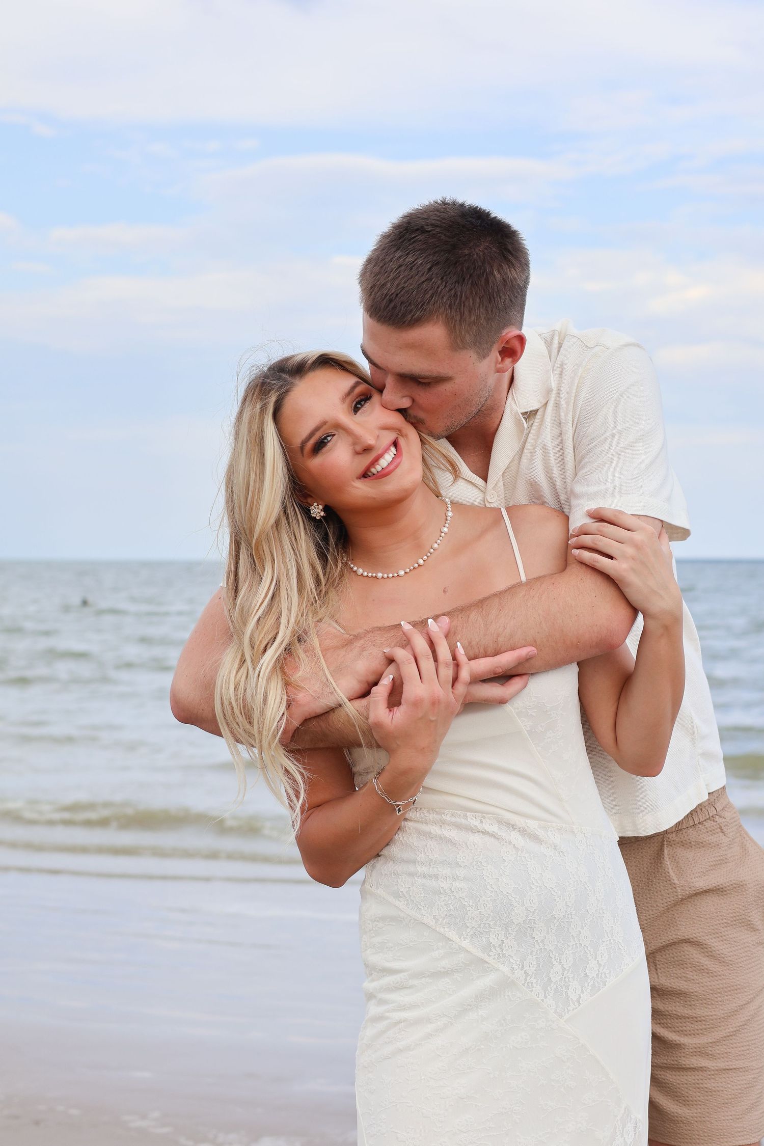 A romantic couple embraces and shares tender moments together on a sunny beach with ocean waves in the background.