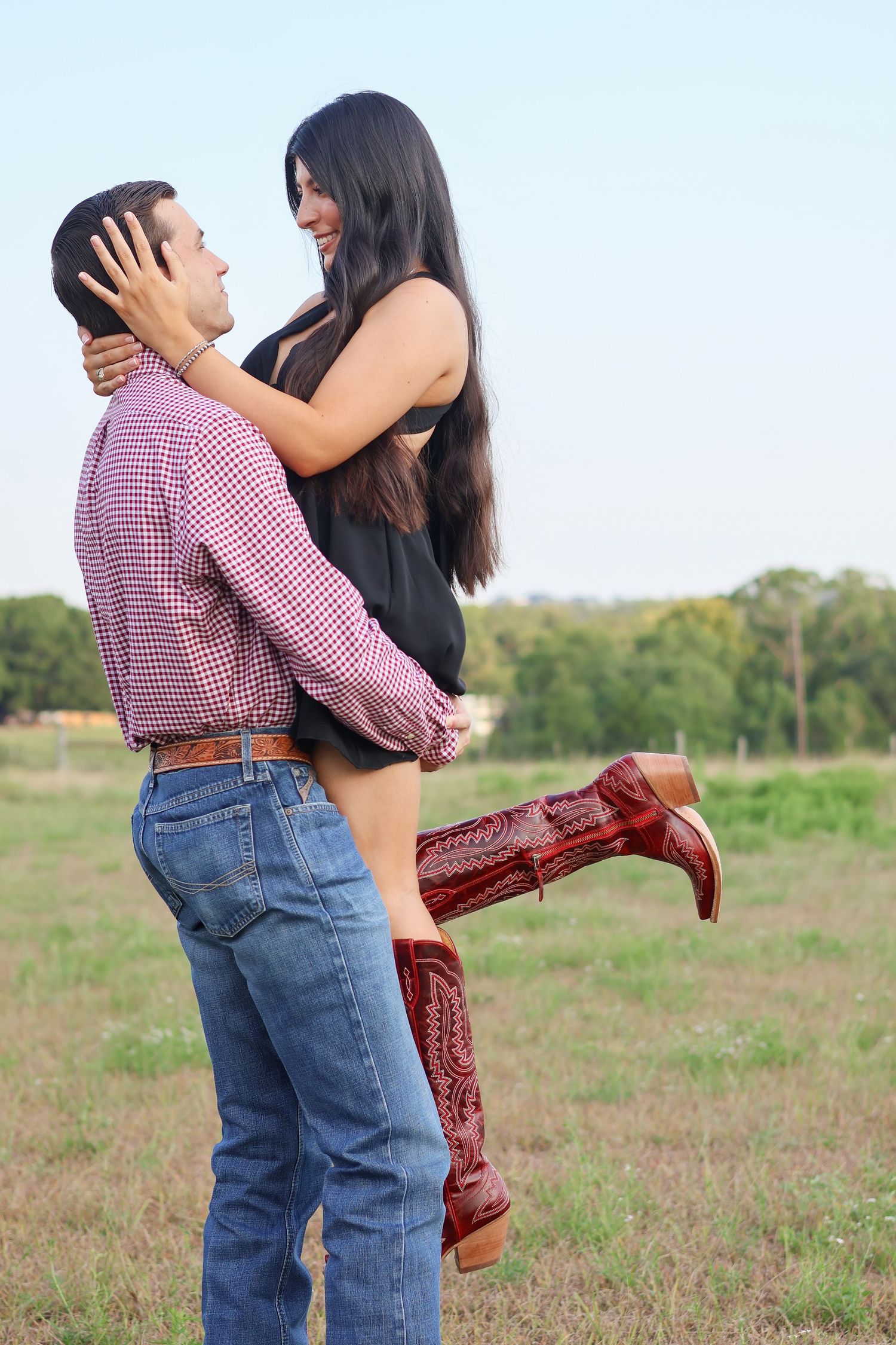 A couple embraces in a field while wearing western attire including red cowboy boots and a plaid shirt.