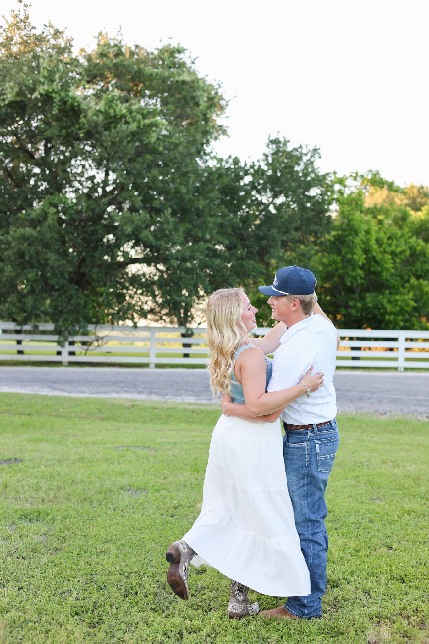 Couple dancing and embracing in a green field with white fence and trees in the background.