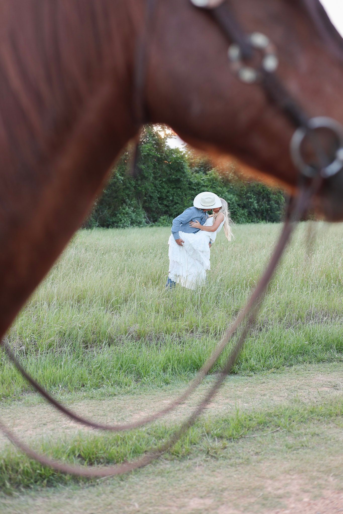 A couple shares a romantic kiss in a green field, photographed through the reins of a chestnut horse.