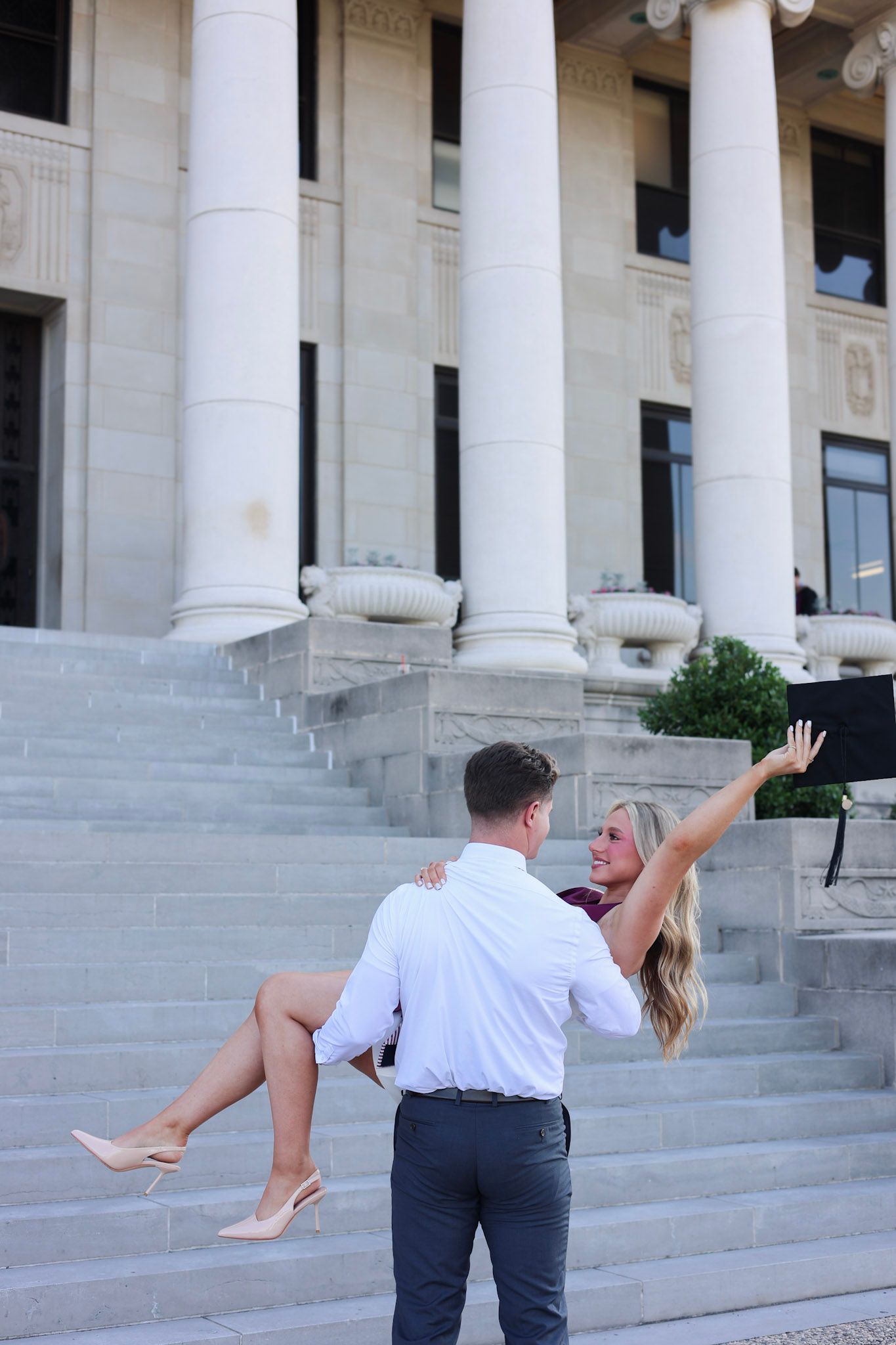 Man carrying woman in his arms on steps of neoclassical building with white columns, woman waving joyfully.