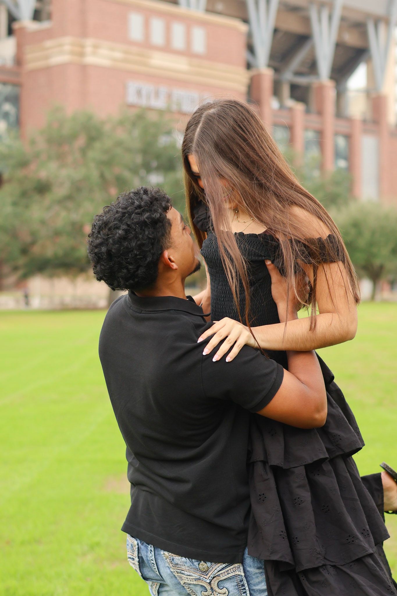 A couple in black outfits share a romantic moment outdoors, with the man on one knee gazing up at the woman.