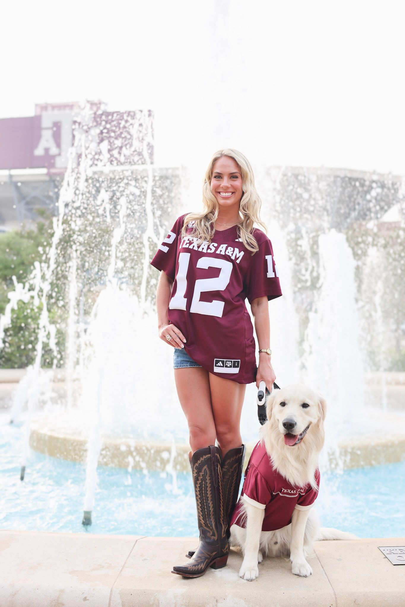 Blonde woman in Texas A&M jersey poses with golden retriever in matching outfit by fountain.