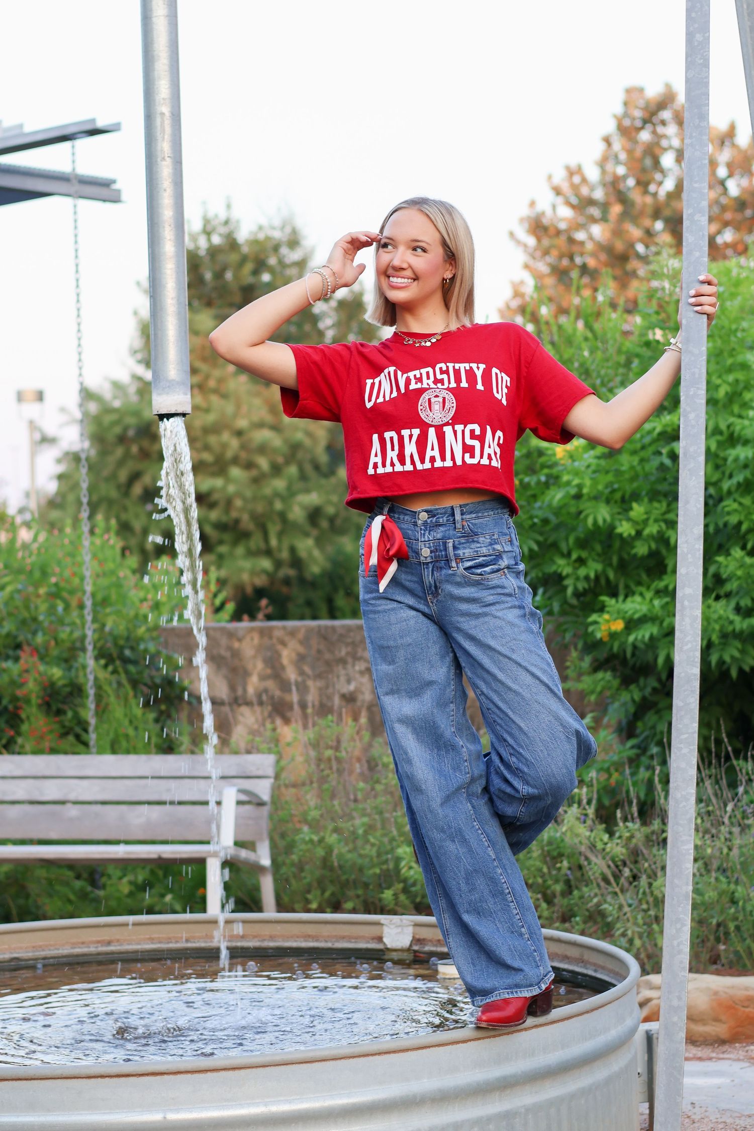 A student in a University of Arkansas crop top and jeans poses casually next to a water tank in an outdoor setting.