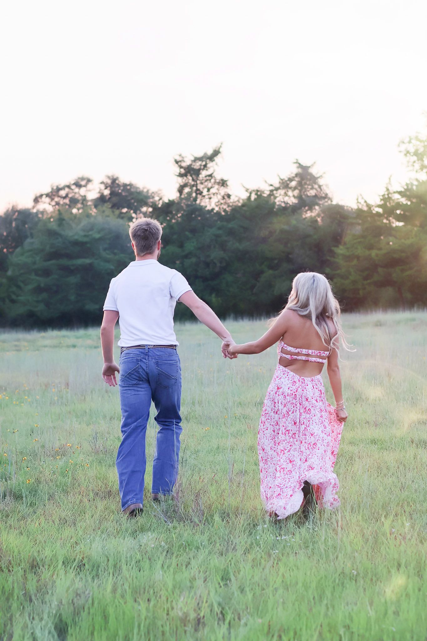 A couple holds hands while walking through a sunlit meadow at golden hour wearing blue jeans and floral dress.