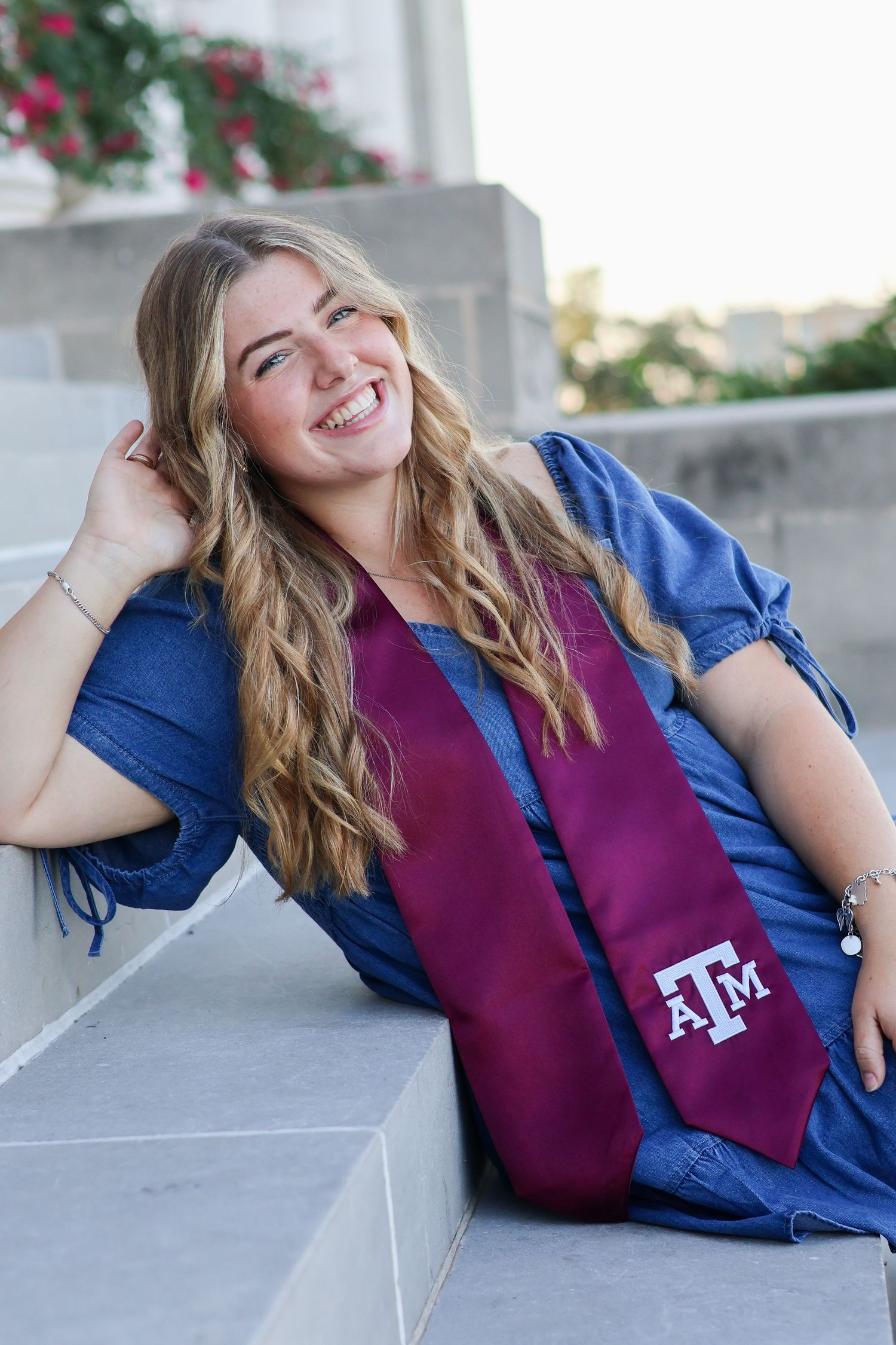 A person in a blue shirt and maroon vest poses for graduation photos while laying on concrete steps.