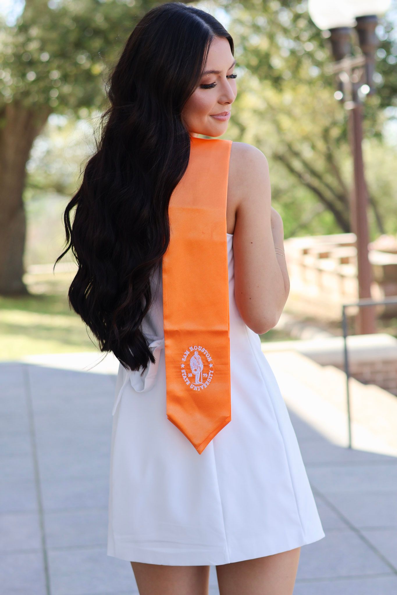 Woman with long dark hair wearing white dress and orange graduation stole standing outdoors on campus.