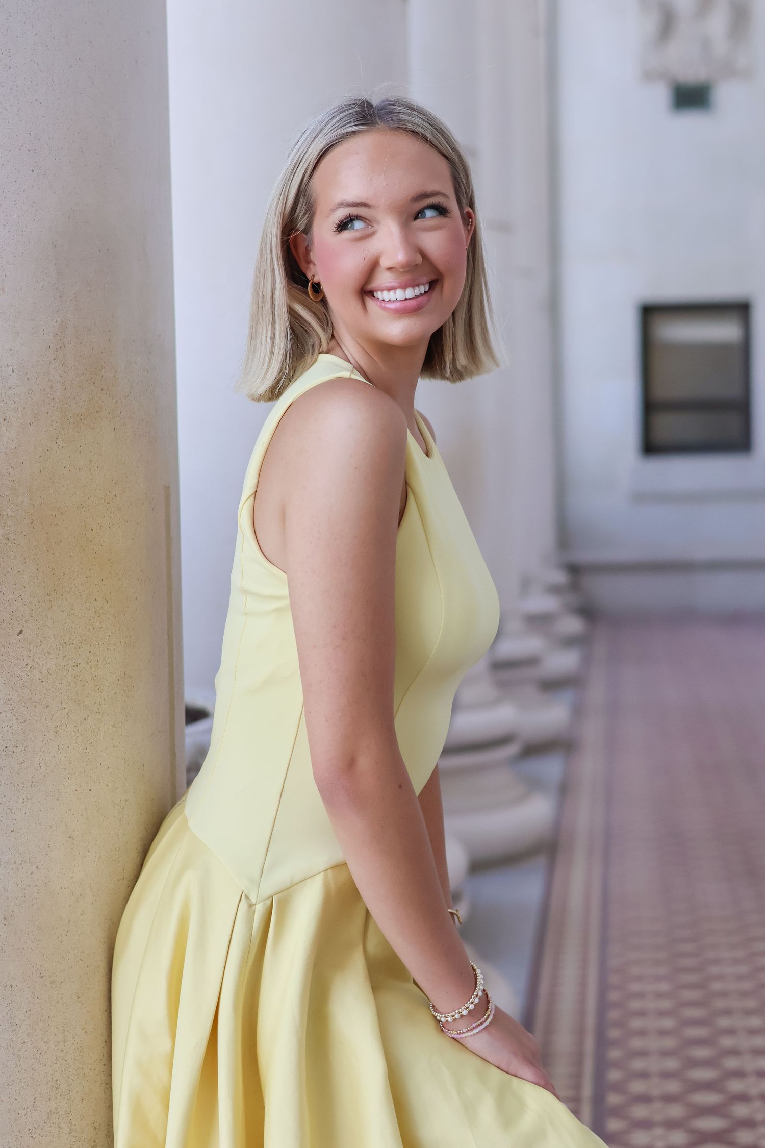Woman in a bright yellow sundress poses and smiles while standing between elegant white columns.