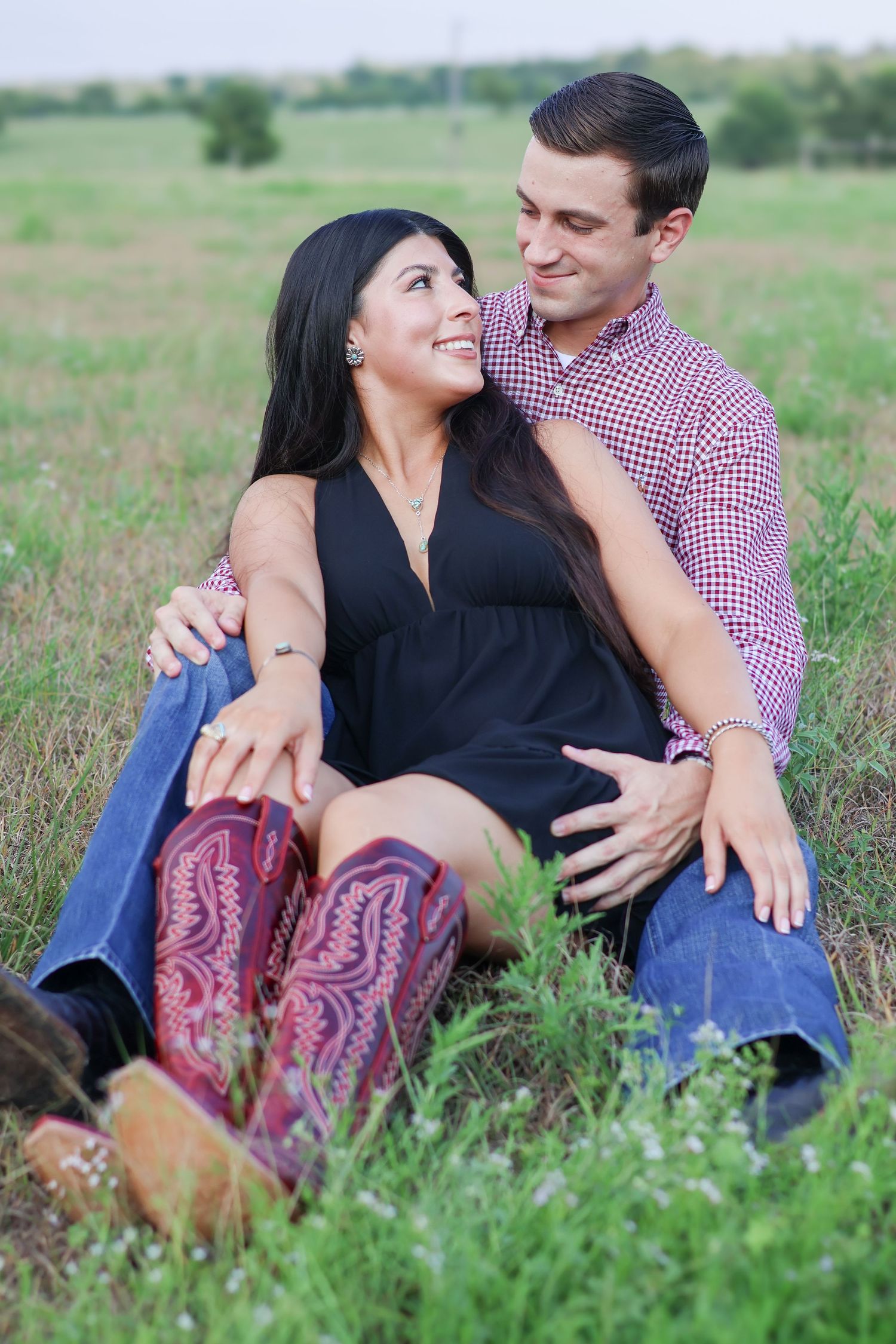 A couple in a red checkered shirt and black dress share intimate moments while sitting together in a grassy meadow.