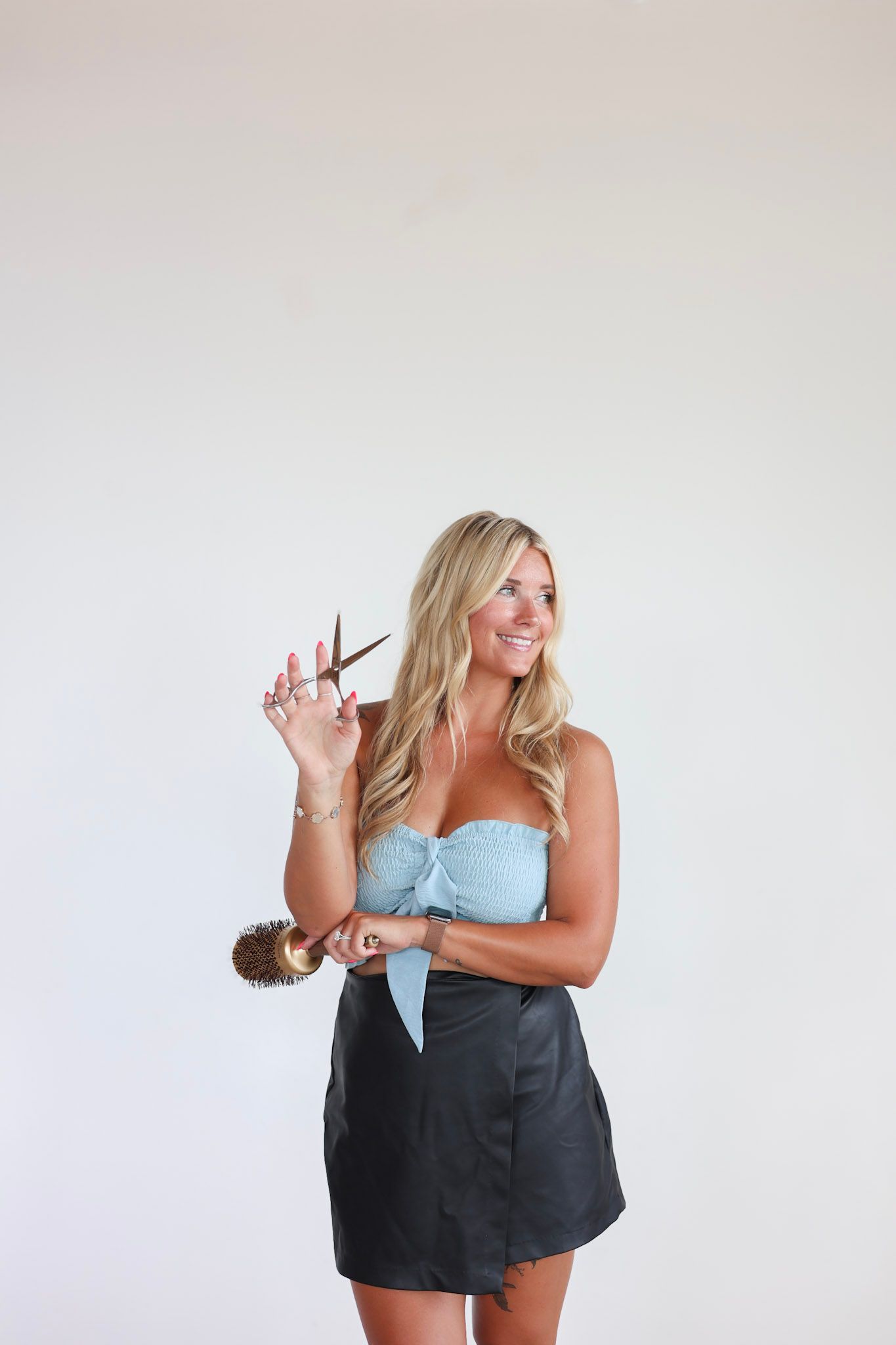 Smiling blonde woman holding hair scissors and a hair tie, wearing a light blue strapless top and black mini skirt.