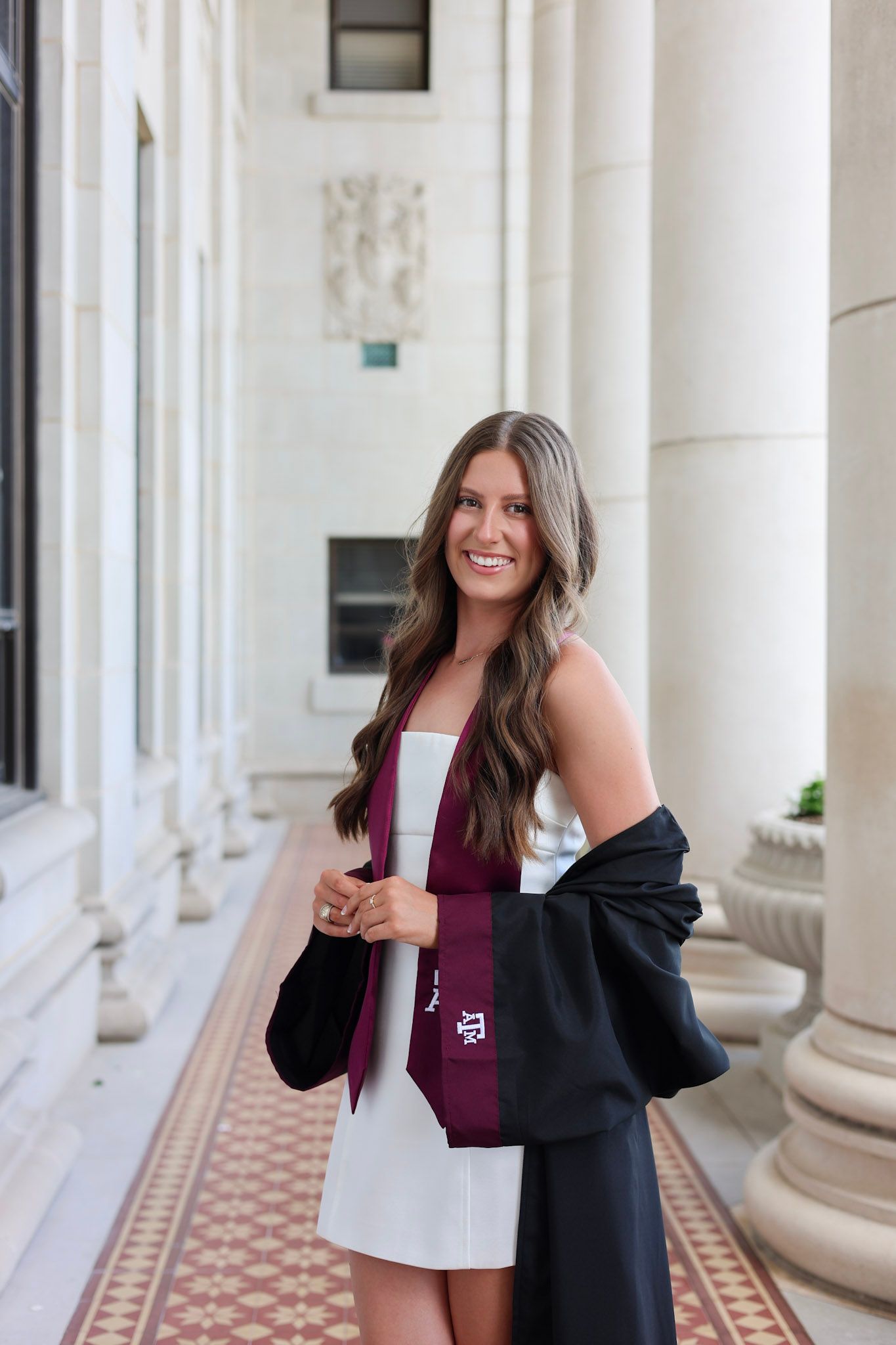 Young woman in white dress holds black graduation gown with Texas A&M maroon sash between columns.