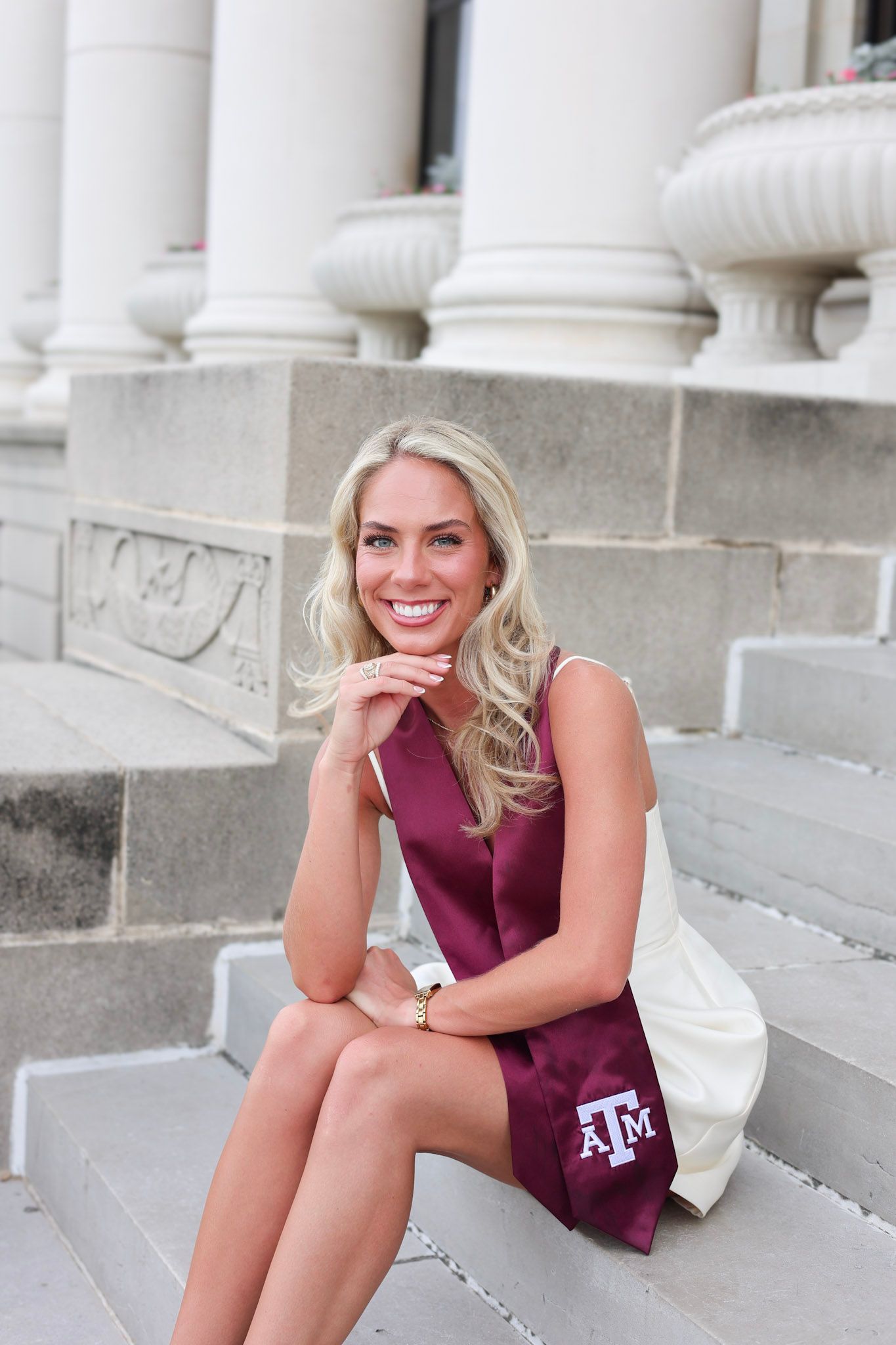 Smiling blonde woman in Texas A&M graduation sash sitting on steps between white columns.