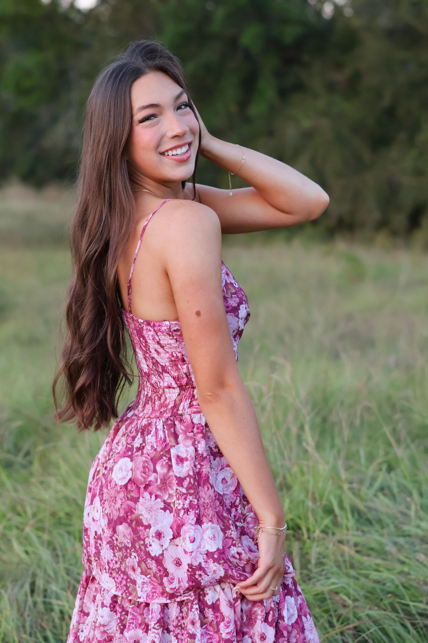 Young woman in pink floral sundress smiling outdoors in a green field, with long brown hair.