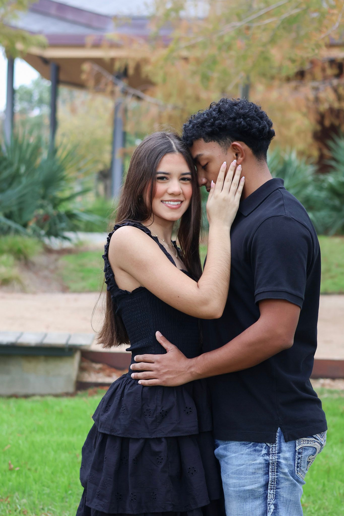 Interracial couple embracing outdoors, woman in black dress smiling at camera while man holds her waist in a park setting.