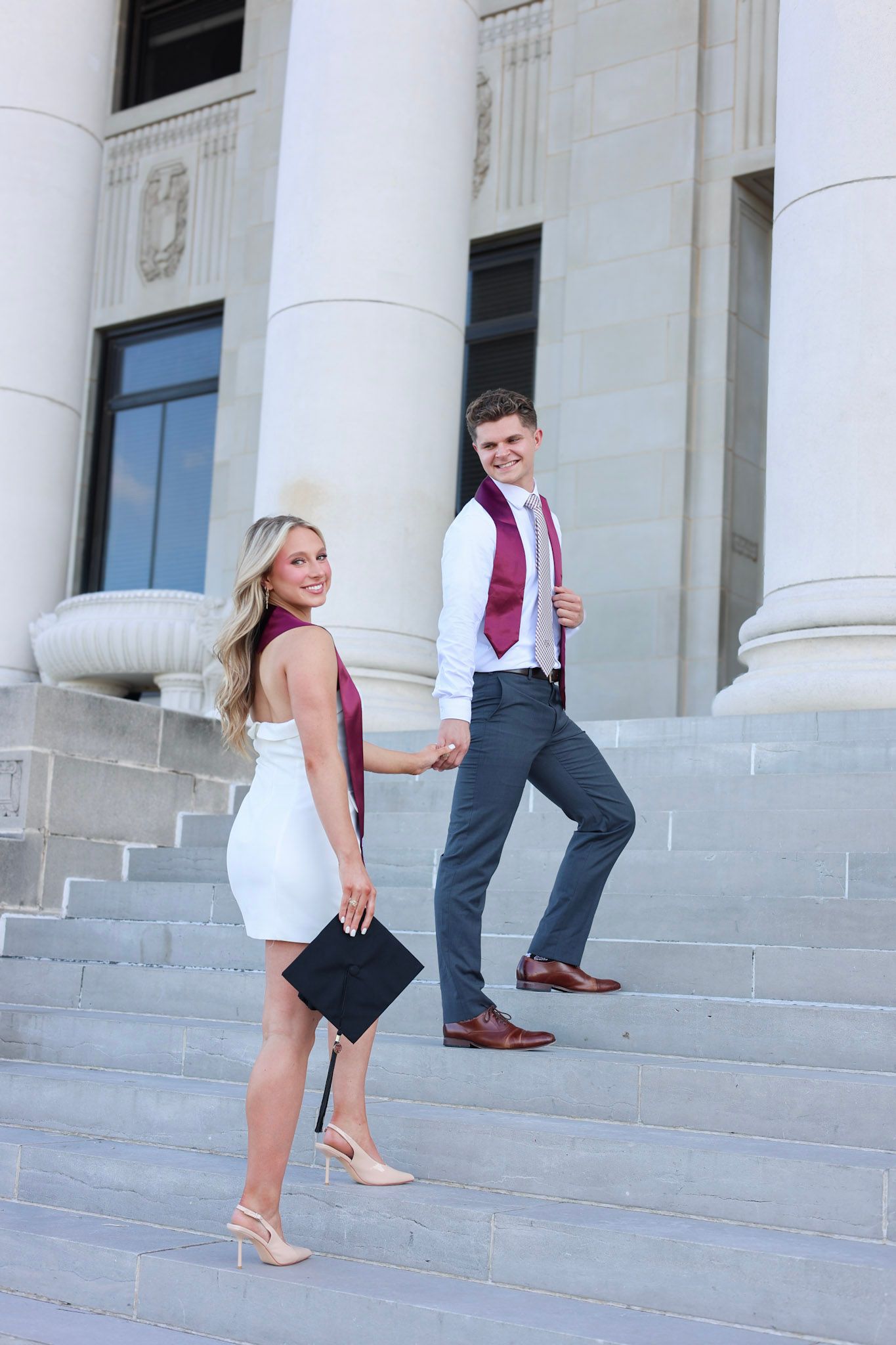 Graduating couple holding hands on steps, woman in white dress holding cap, man in maroon tie.