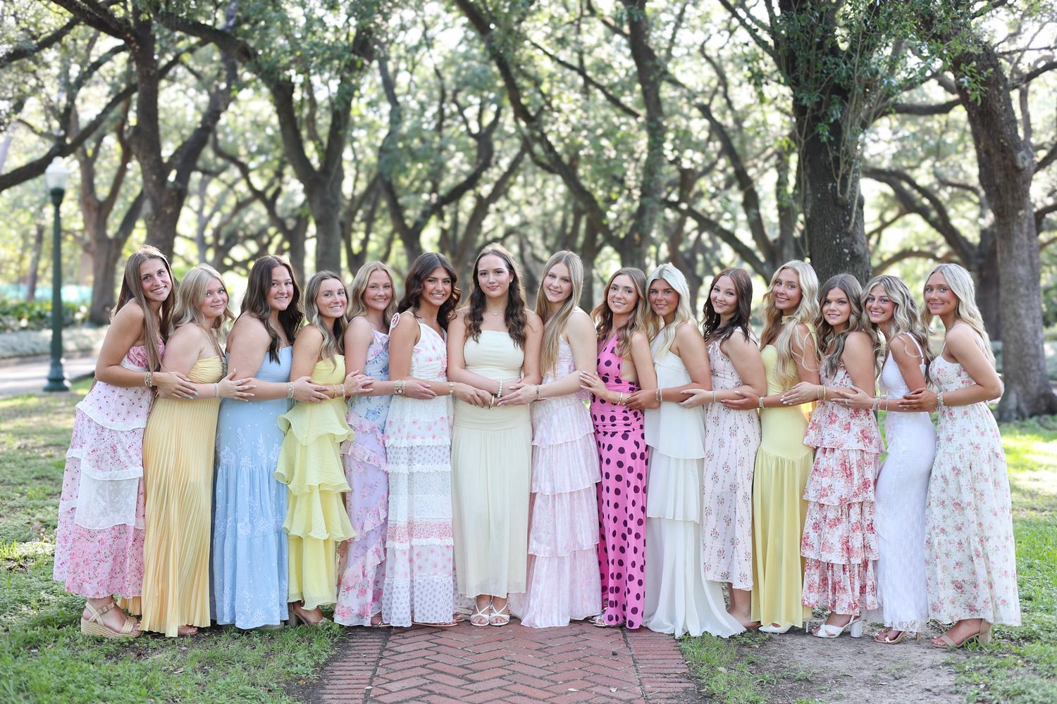 Group of young women in pastel maxi dresses posing together outdoors under oak trees on a brick path.