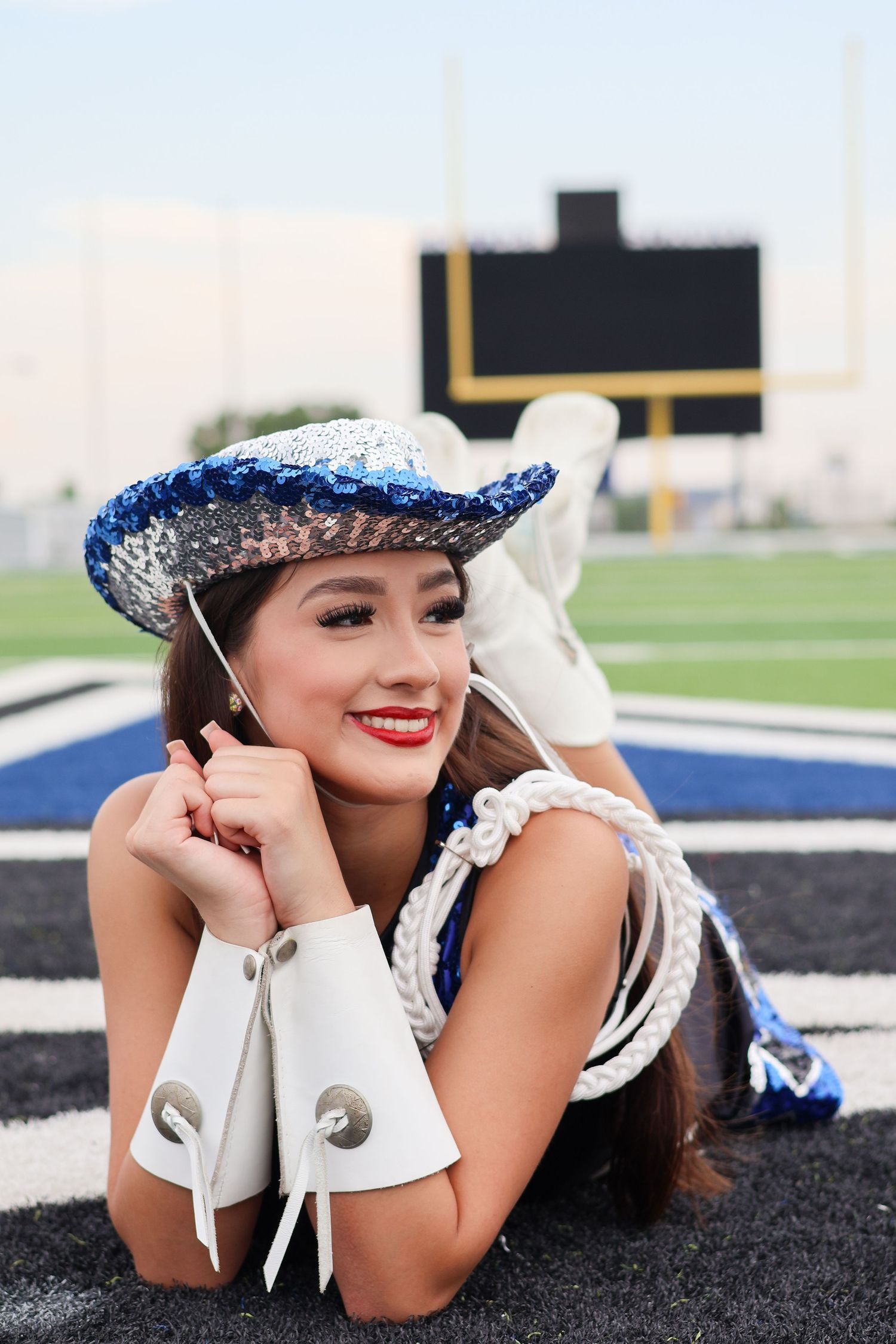 A cheerleader in blue and white uniform with sparkly hat lies on a football field with goal post visible in background.