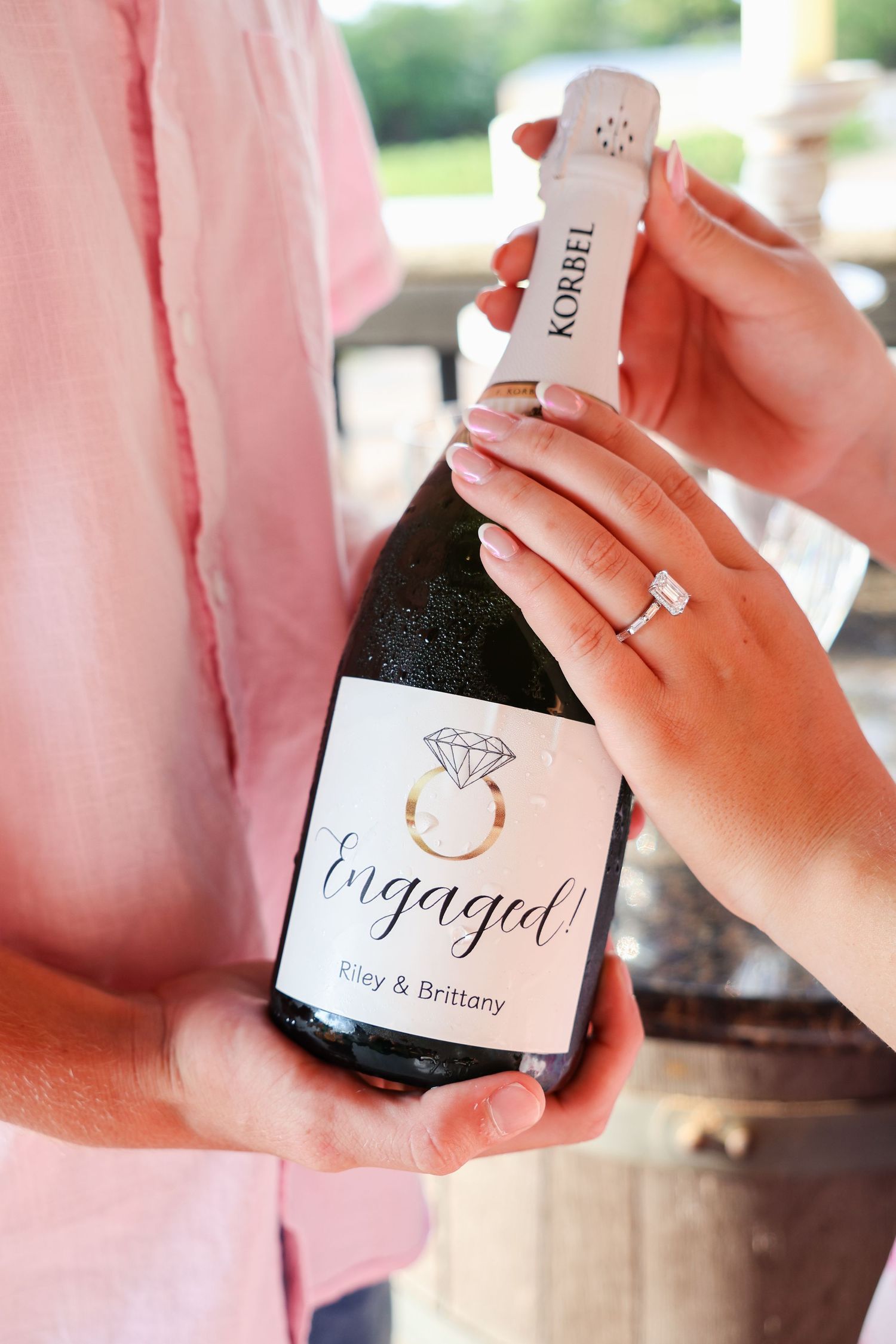 Wine bottle with 'Engaged' label being held against pink background showing romantic proposal announcement.