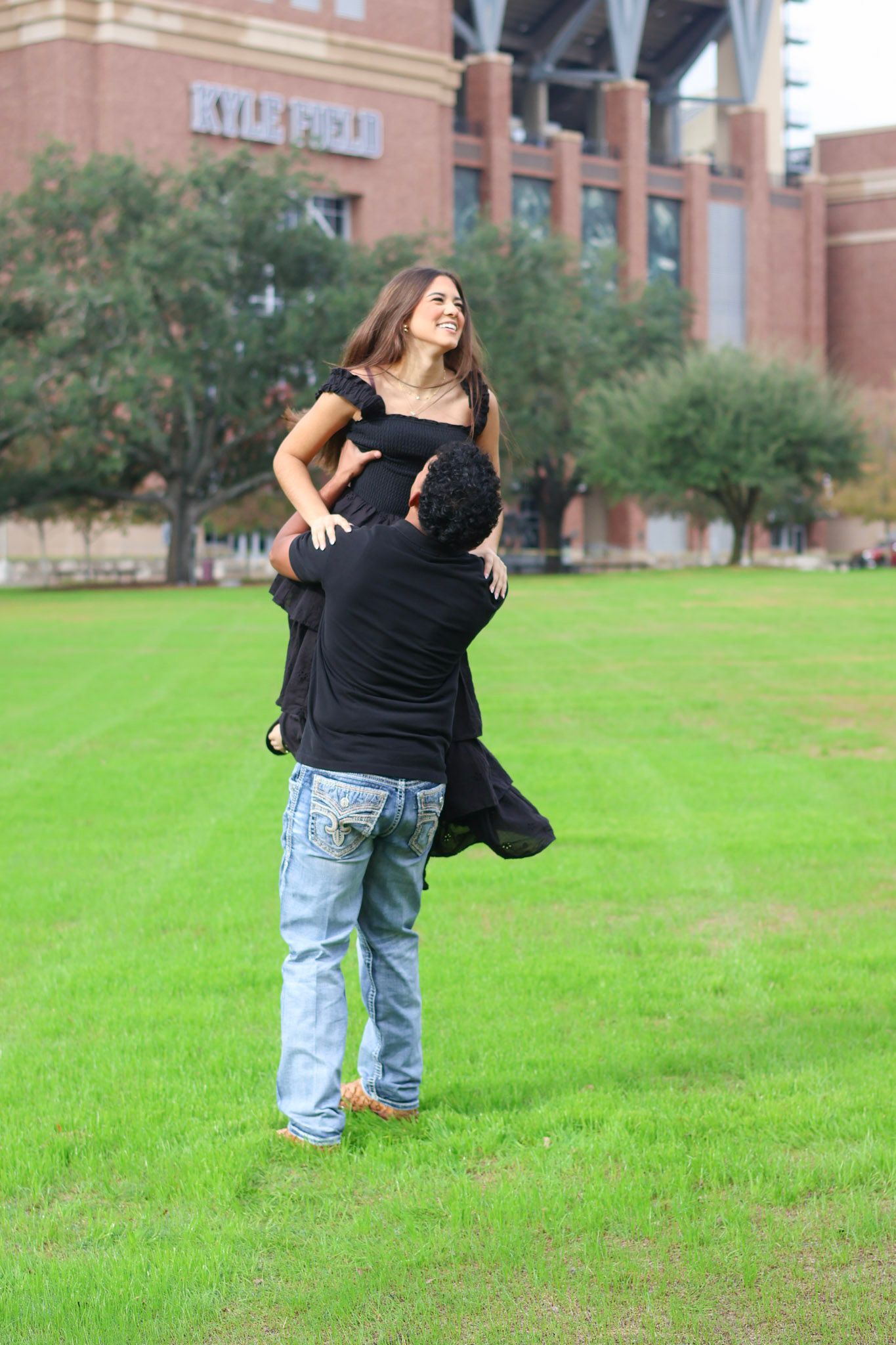 Man in jeans lifting woman in black dress outdoors near Kyle Field stadium on a green lawn.
