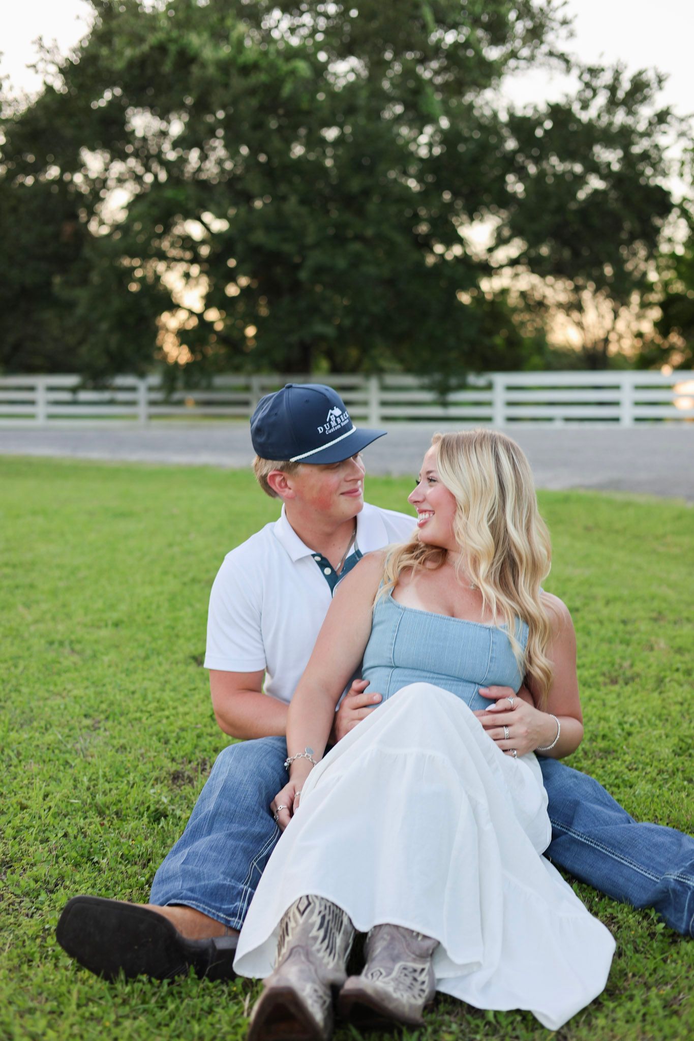 A couple sits on green grass, smiling at each other, man in white polo and cap, woman in blue dress with cowboy boots.