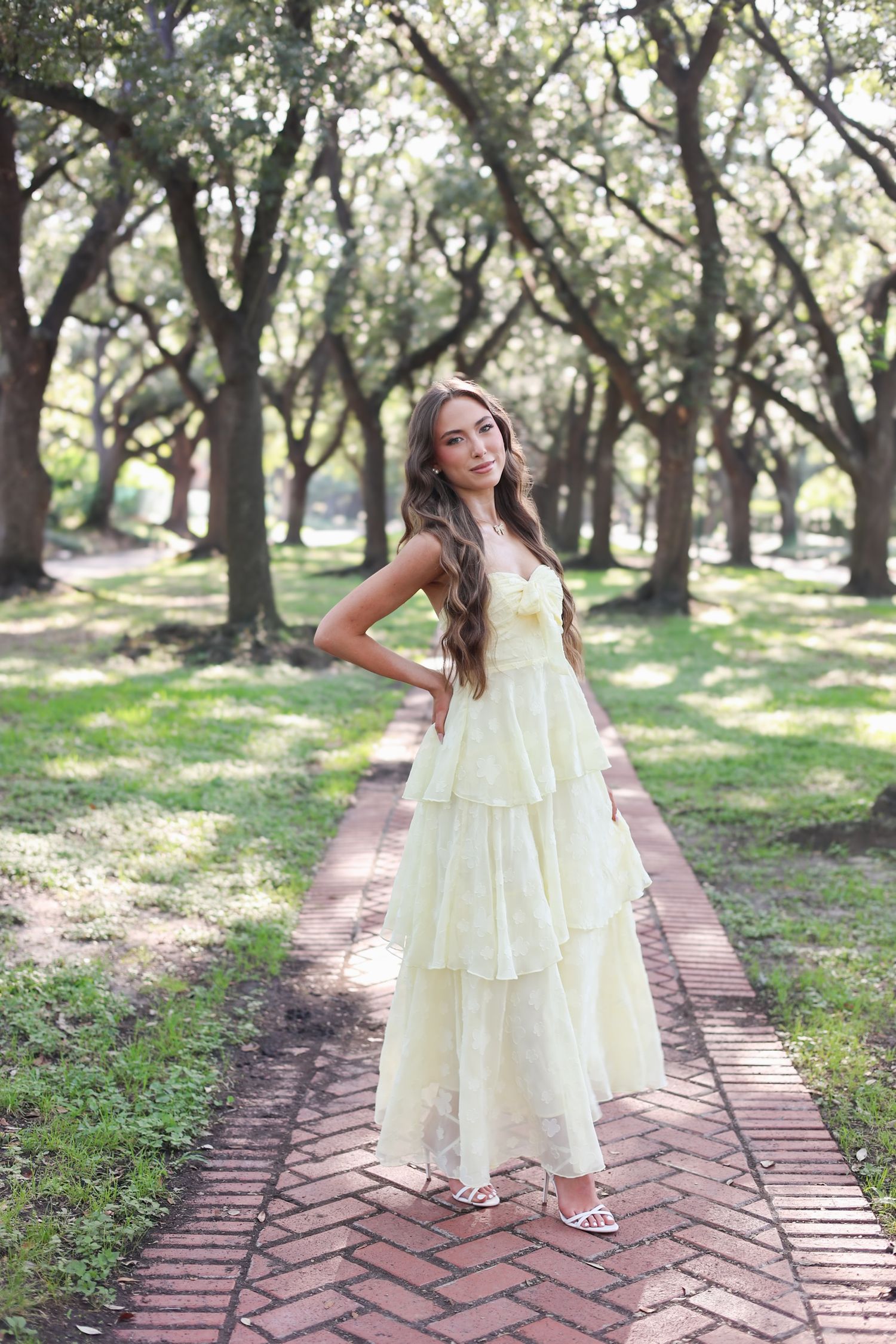 Young woman in a flowing yellow tiered dress poses on a brick path surrounded by oak trees.