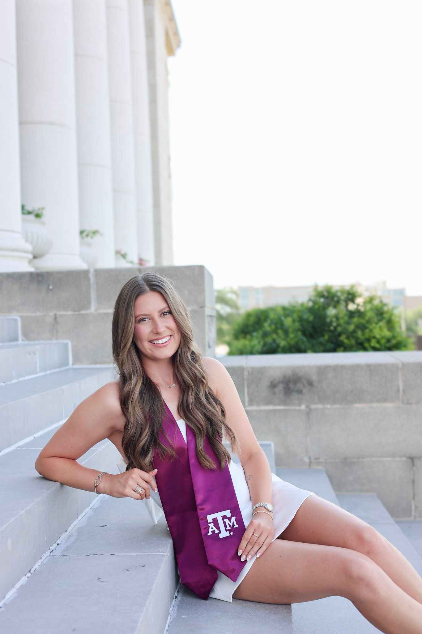 Texas A&M graduate smiling in white dress and maroon stole seated on university steps.