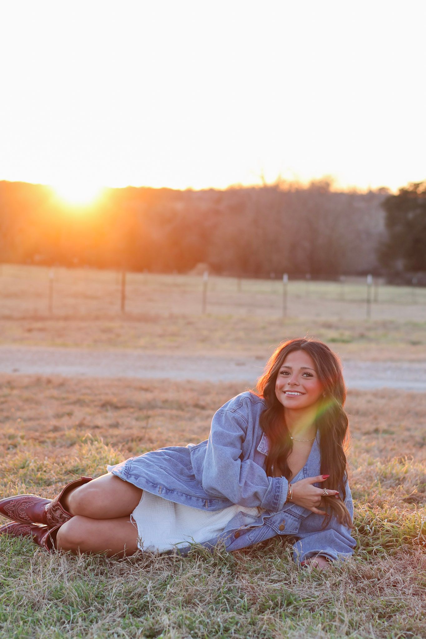 Young woman in denim jacket and cowboy boots sitting in a field at golden hour sunset.