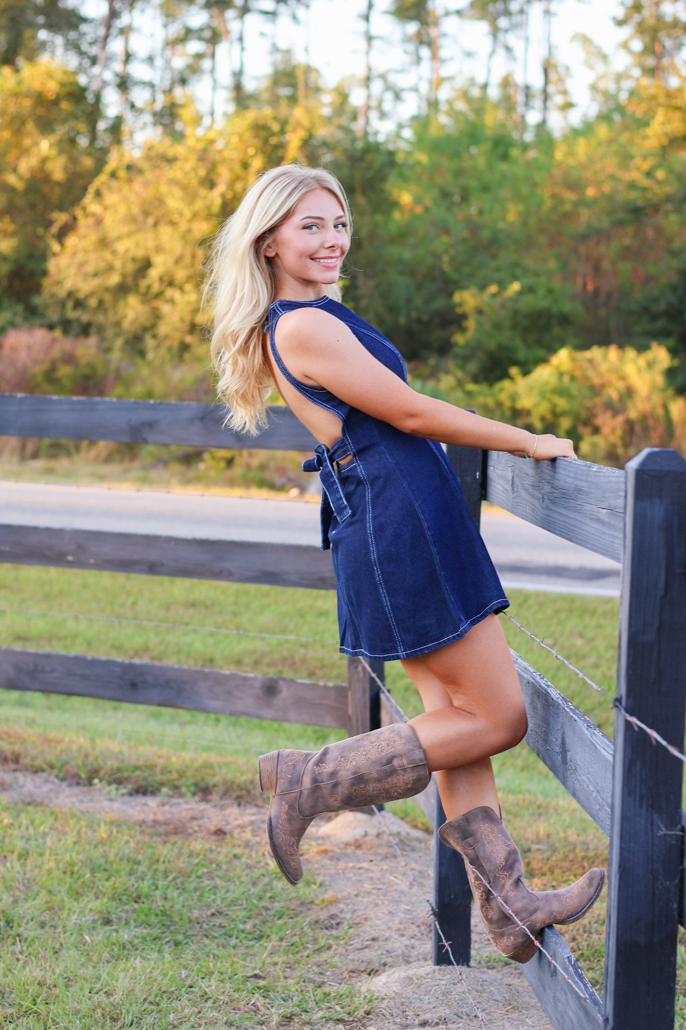 Smiling blonde woman in blue denim dress and cowboy boots posing by a wooden fence outdoors.