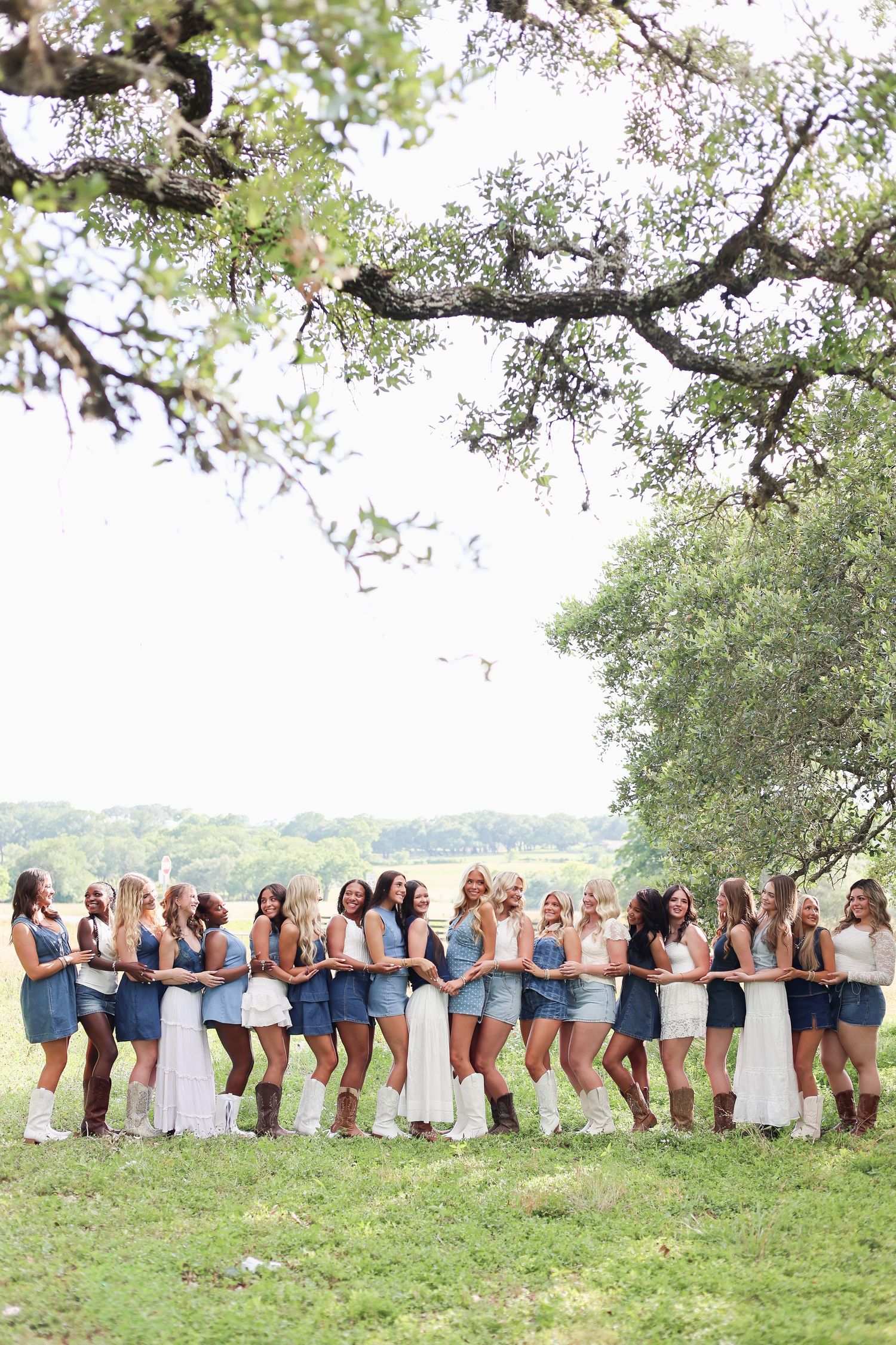 Large bridal party in denim outfits and cowboy boots posing under oak trees at an outdoor country wedding.