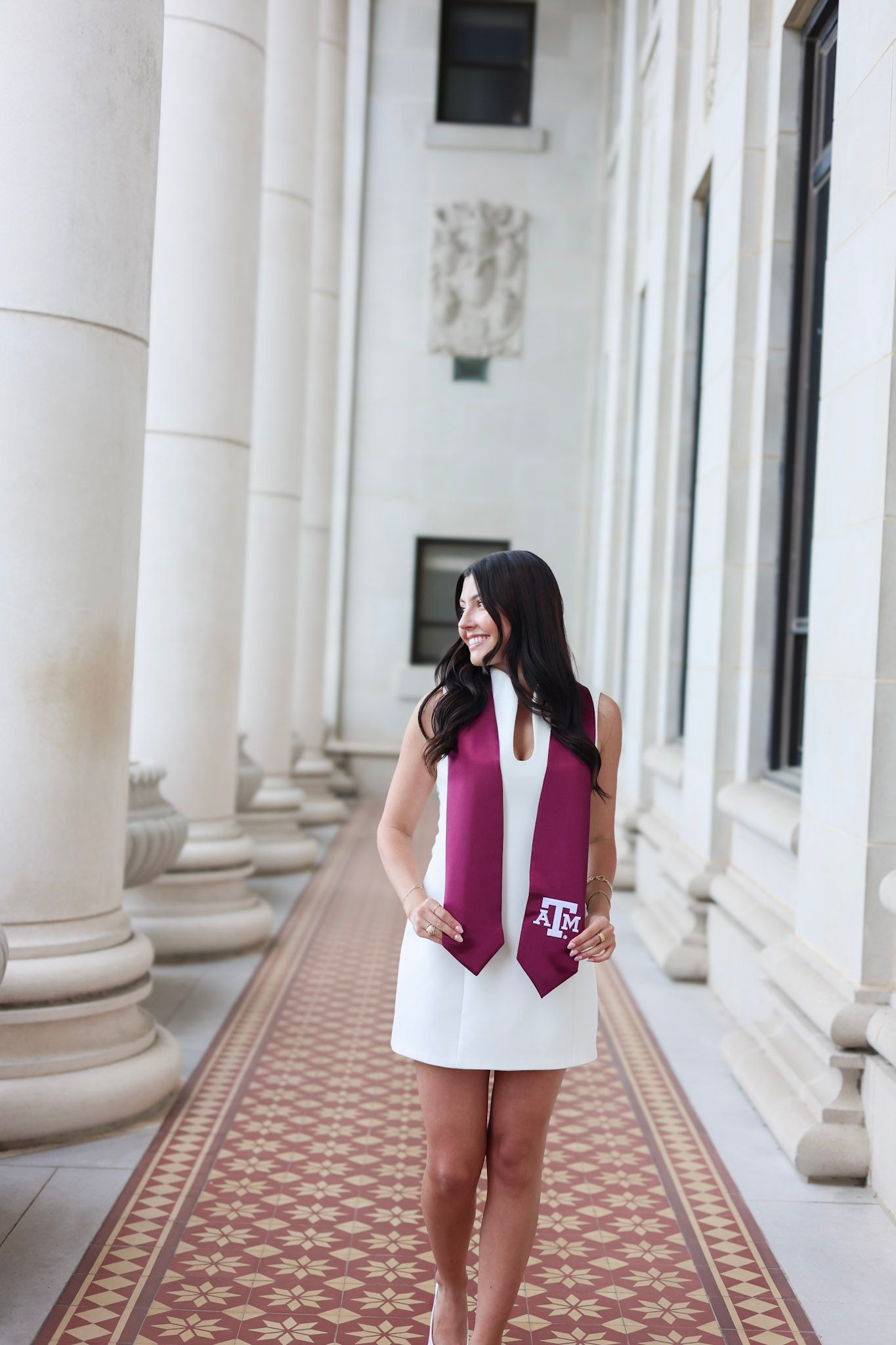 Young woman in white dress wearing Texas A&M maroon graduation stole, smiling in columned hallway.