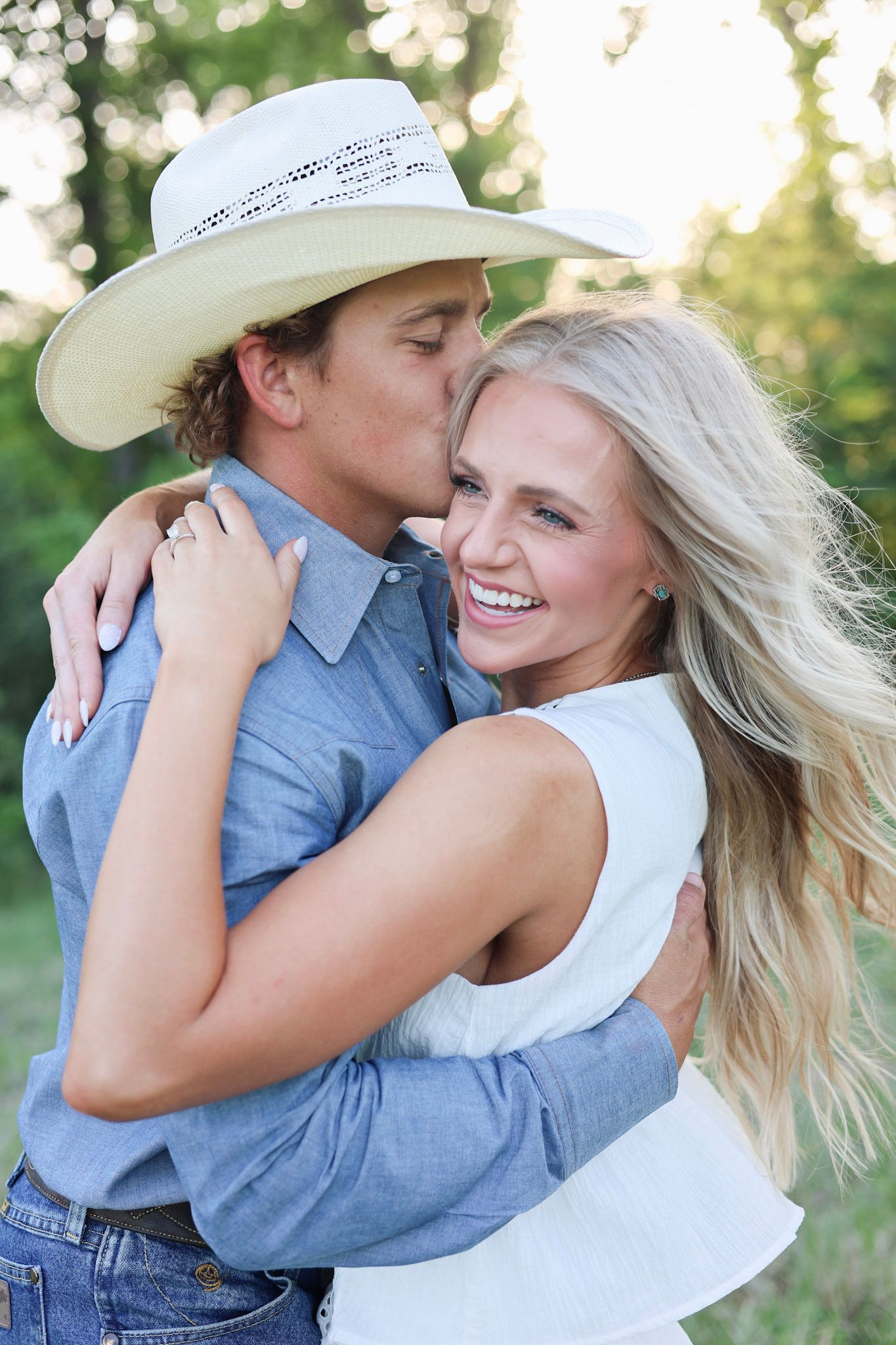 A smiling blonde woman hugs a man in a cowboy hat and blue shirt outdoors in a romantic couples portrait.
