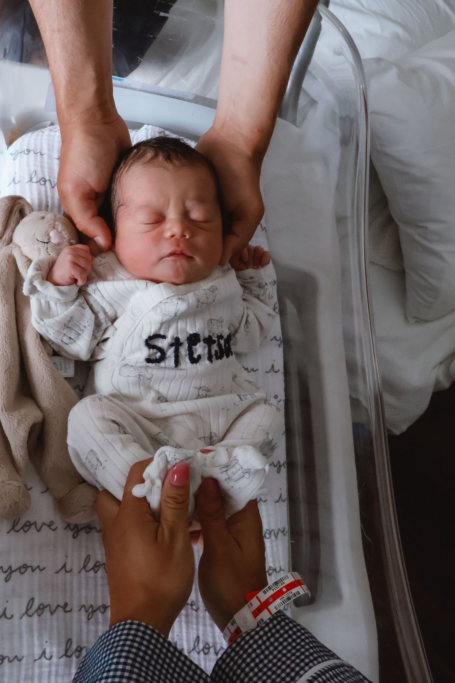 Newborn baby wrapped in text-printed blanket being held gently during hospital photoshoot.