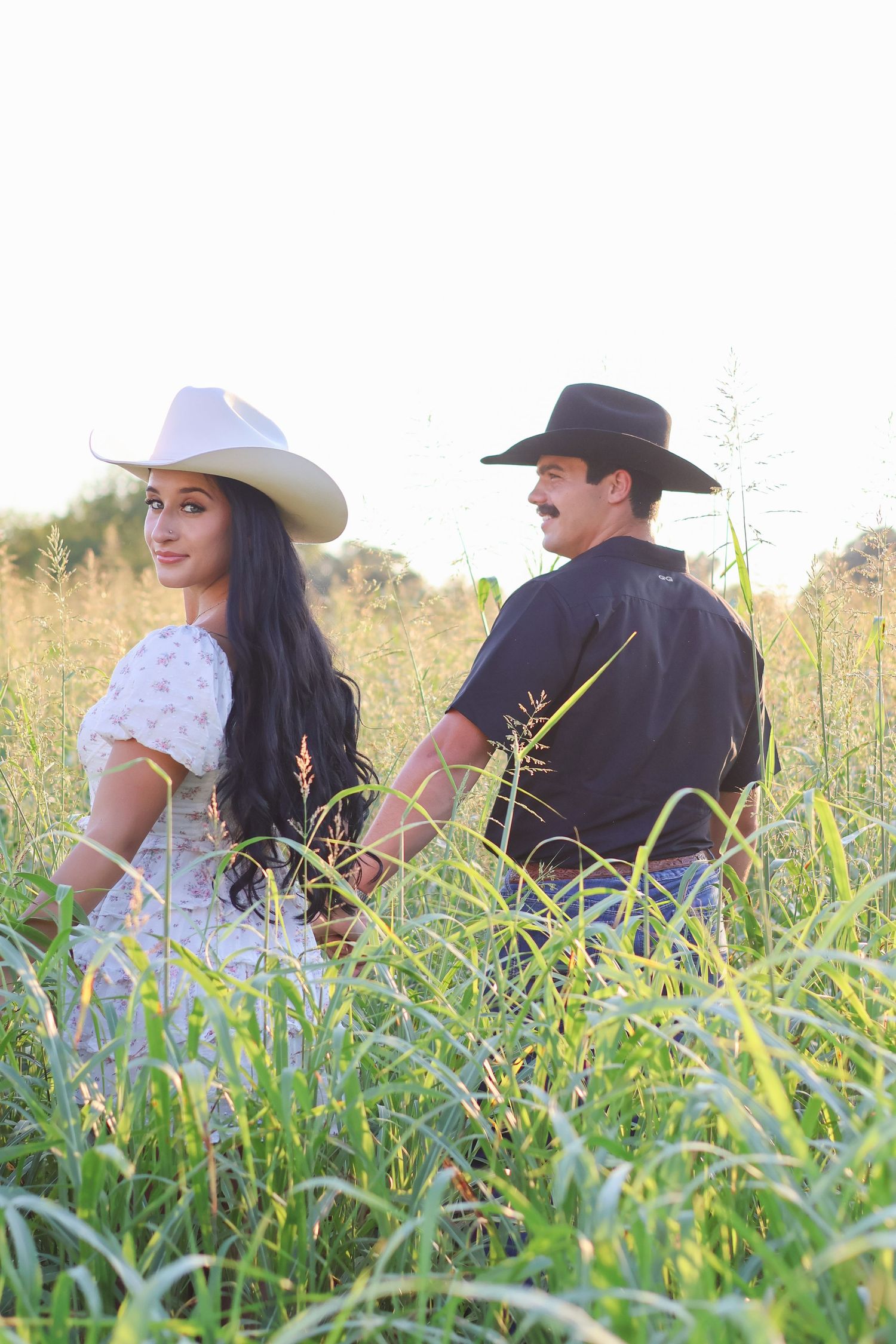 A couple in western attire sits together in a tall grassy field during golden hour.