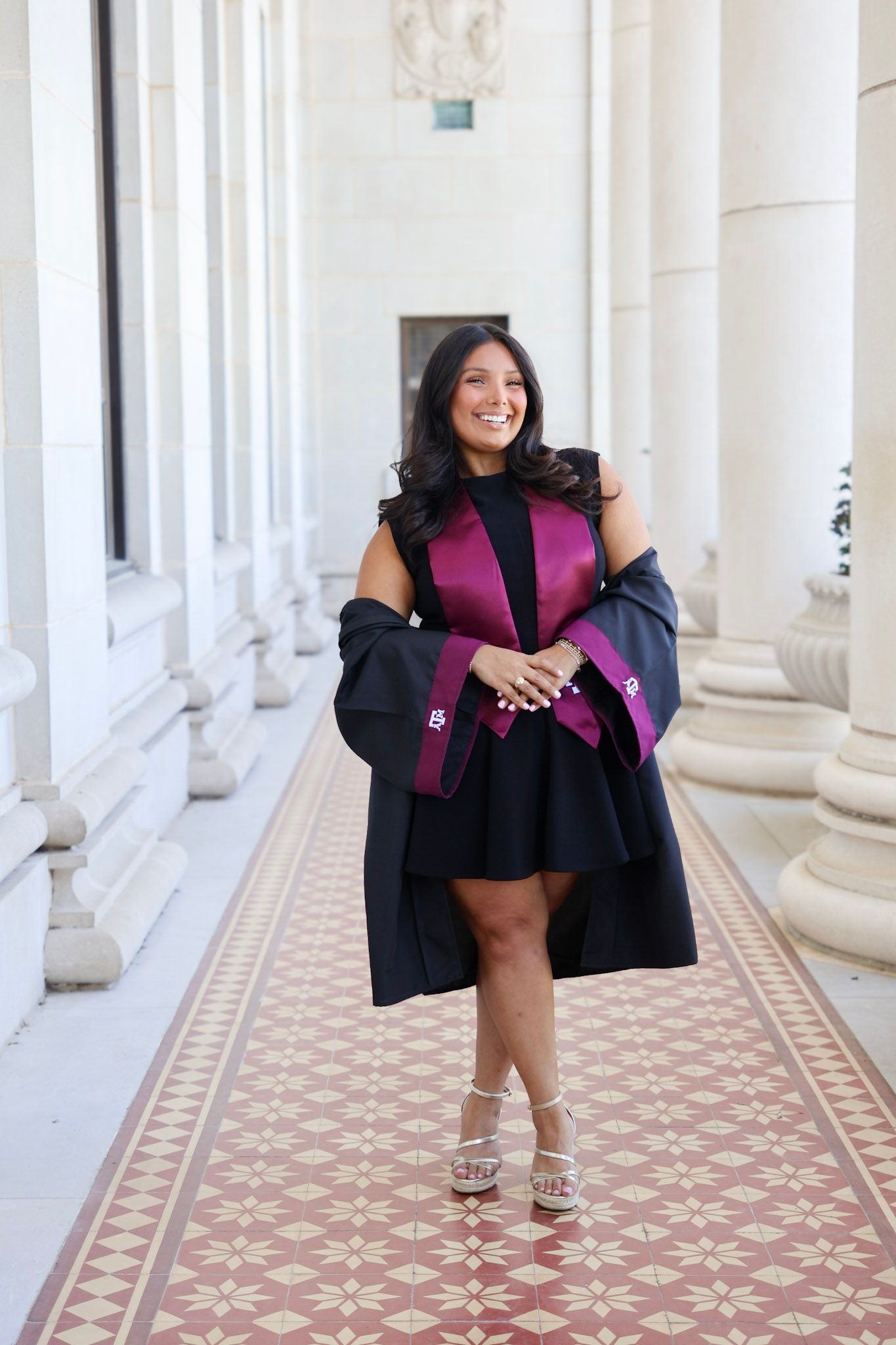 Smiling graduate in black gown with purple satin trim poses in elegant columned hallway with mosaic tile floor.