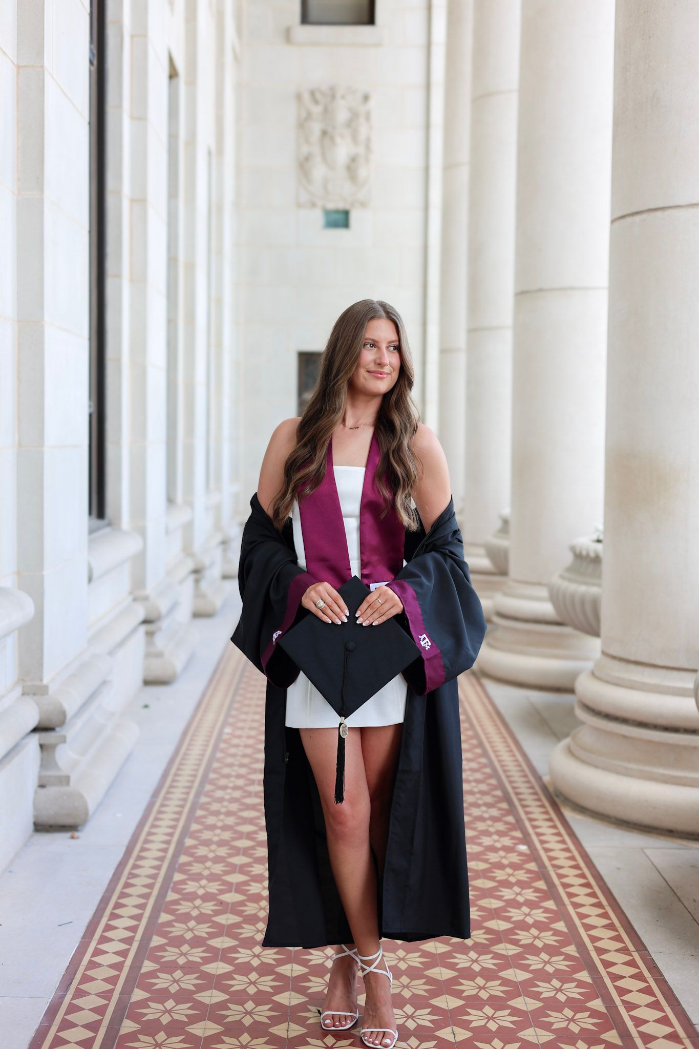 Texas A&M senior holds graduation cap while wearing black gown and maroon stole between elegant white columns.